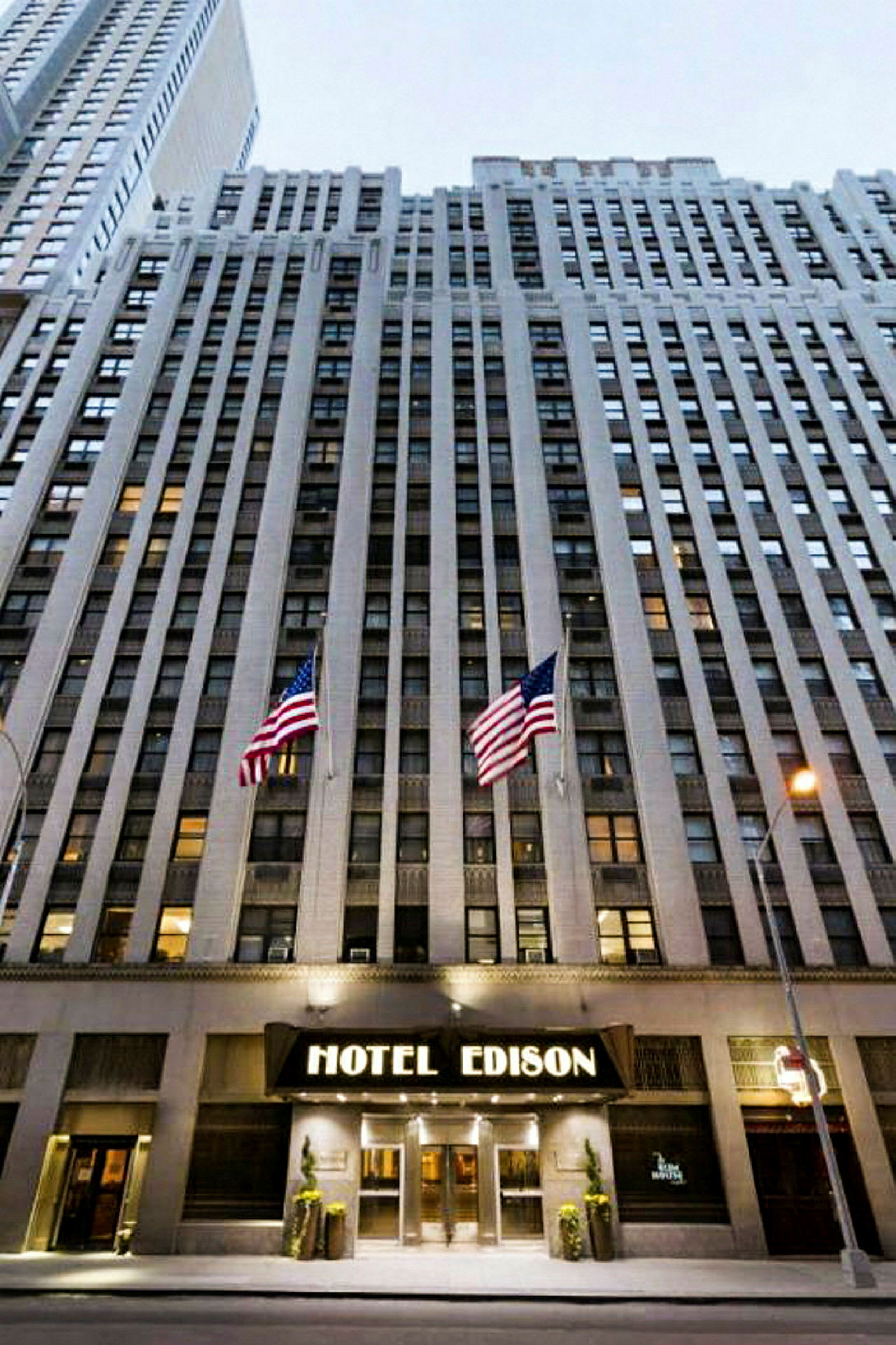 Udsigt til en høj bygning med indgangen markeret "Hotel Edison" og to amerikanske flag foran. (AI-genereret tekst)