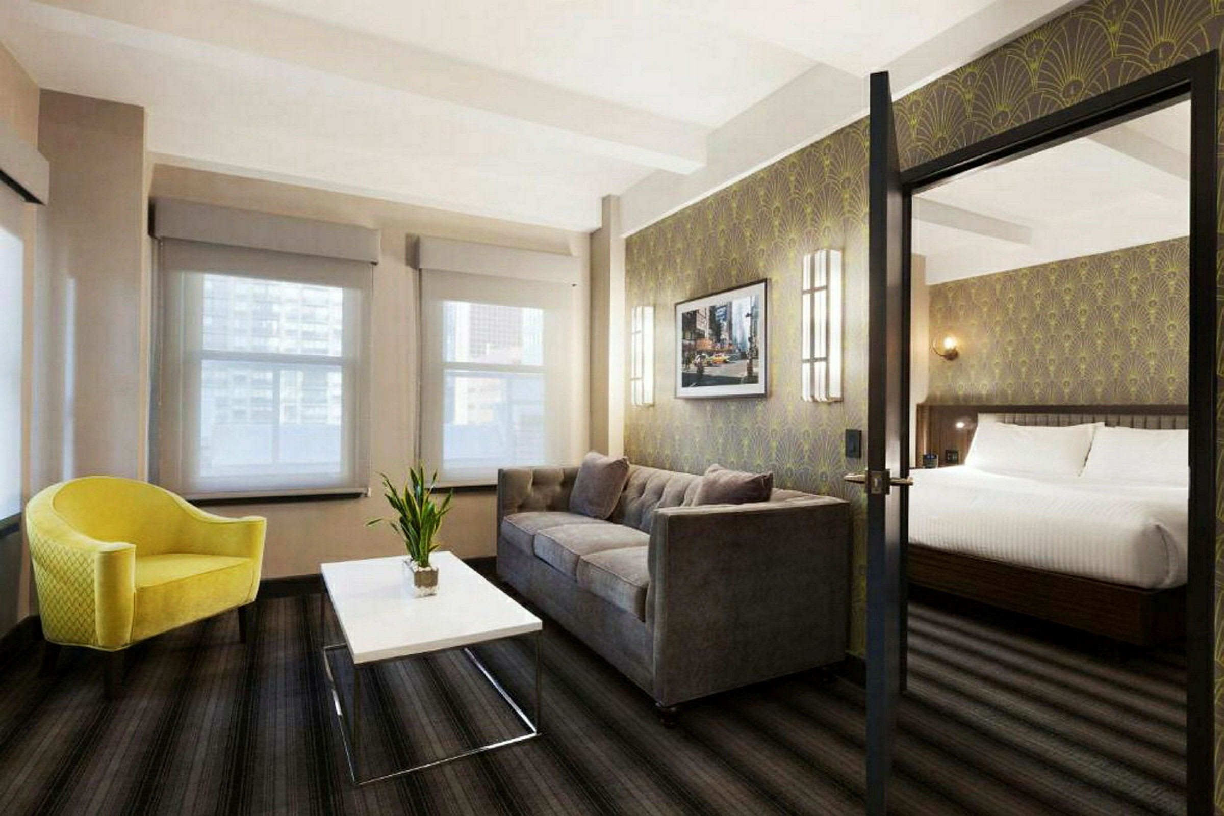 Et hotelværelse med en siddegruppe bestående af en gul lænestol, et hvidt sofabord og en grå sofa. I baggrunden ses en soveplads med en dobbeltseng. (AI-genereret tekst)