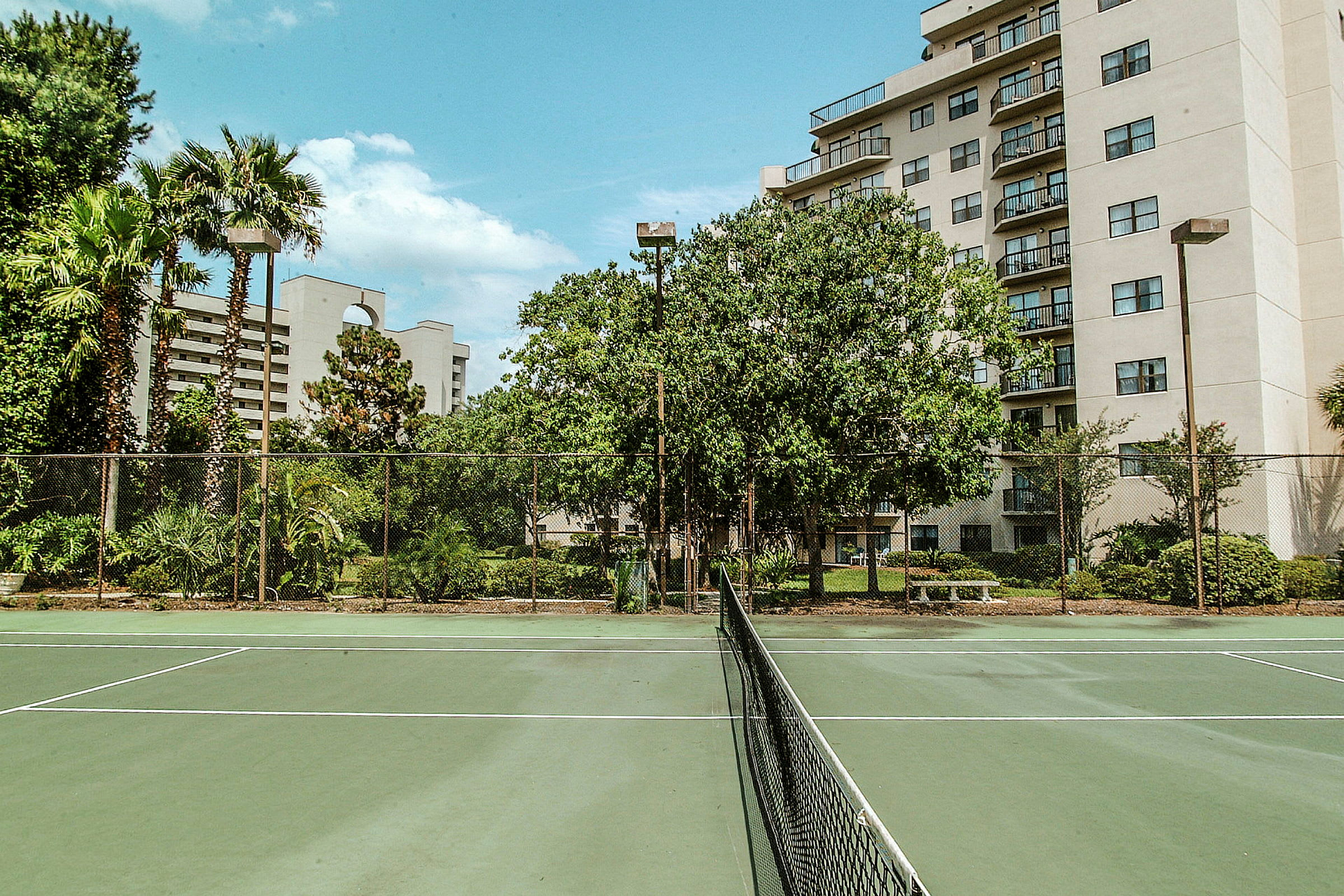Tennisbane omgivet af grønne planter og bygninger i baggrunden. (AI-genereret tekst)