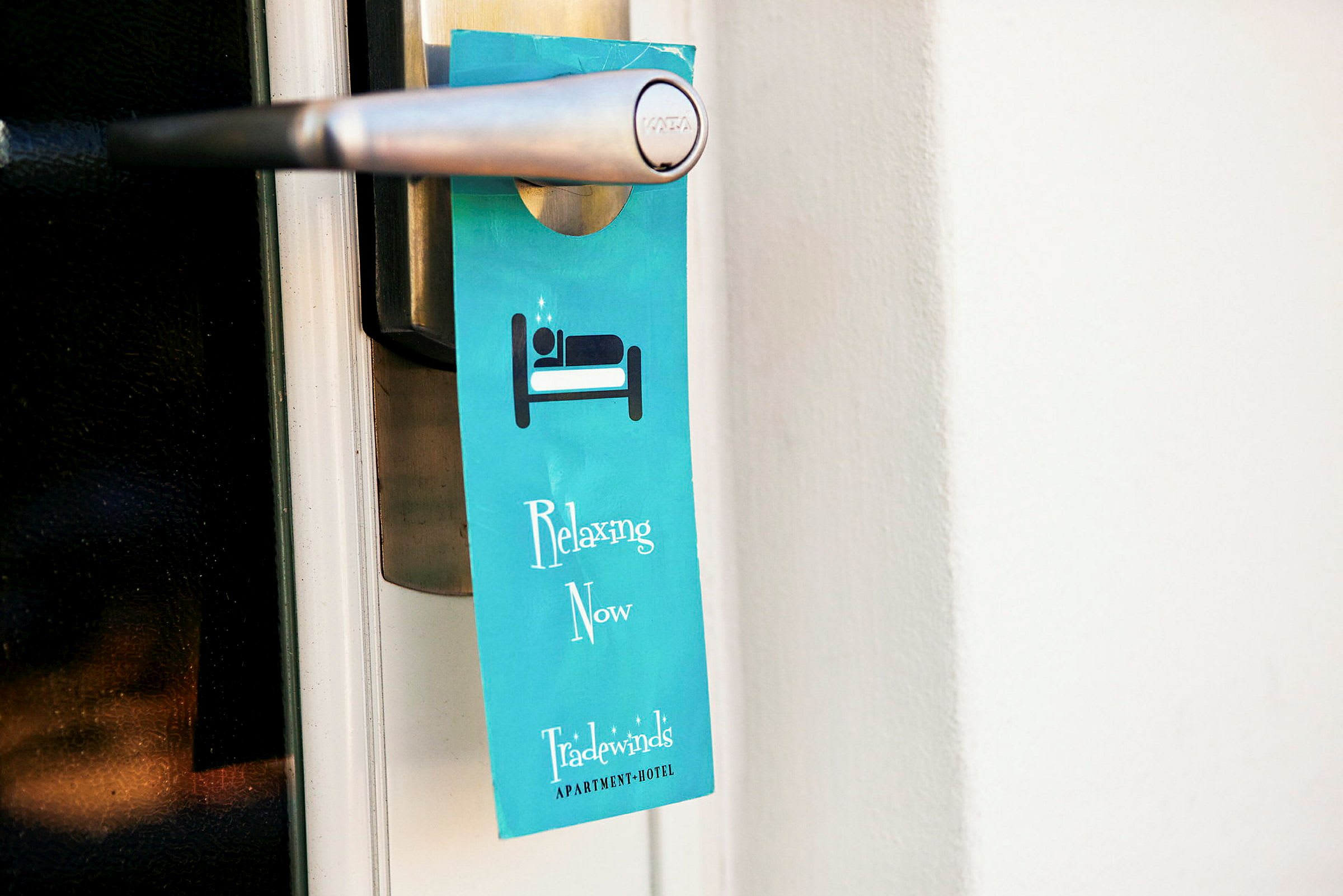 En blå skilt hænger på et dørhåndtag med teksten "Relaxing Now" og hotellets navn. (AI-genereret tekst)