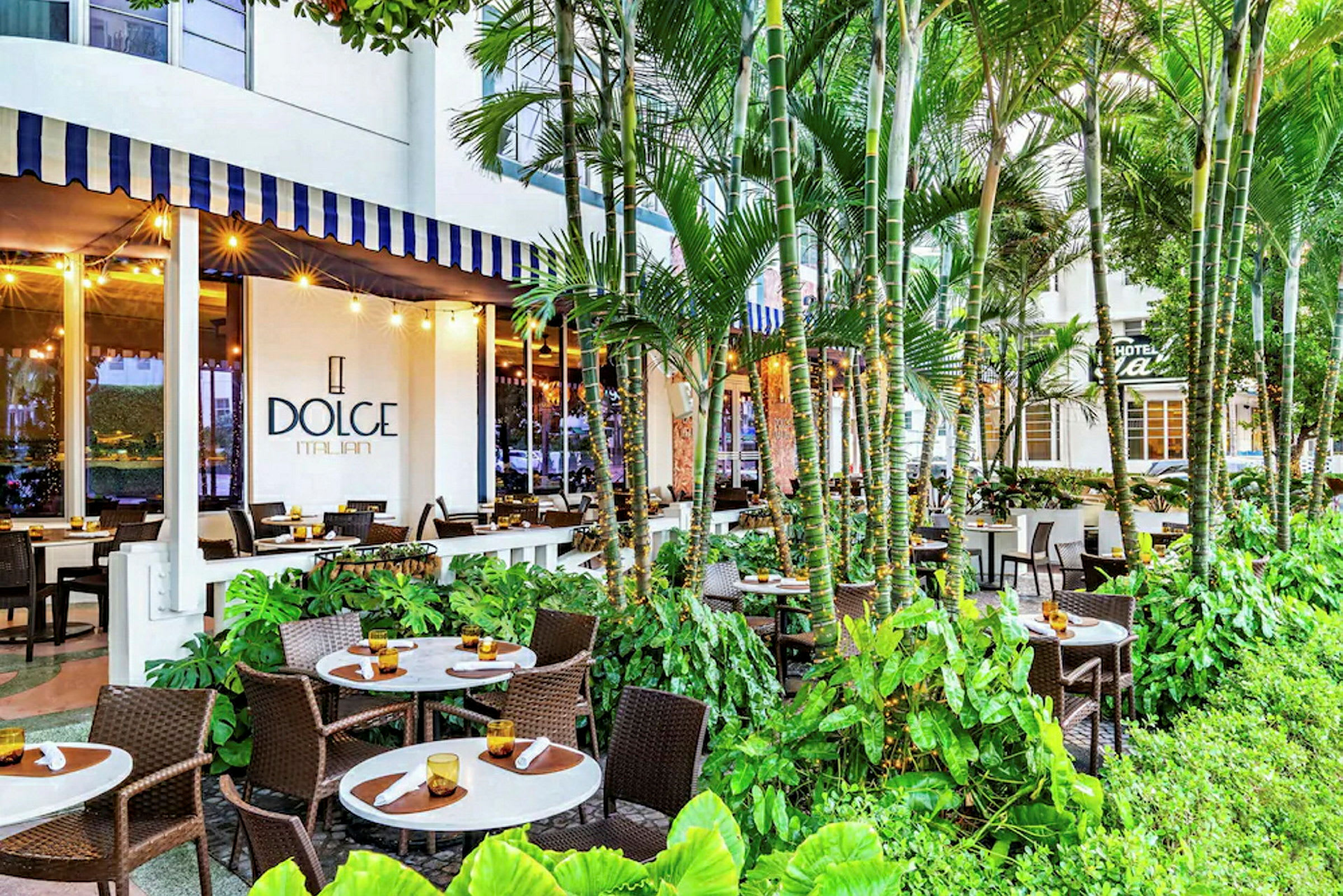En udendørs restaurant med flere borde og stole omgivet af frodigt grønt, med et skilt for 'Dolce Italian' synligt på bygningen. (AI-genereret tekst)
