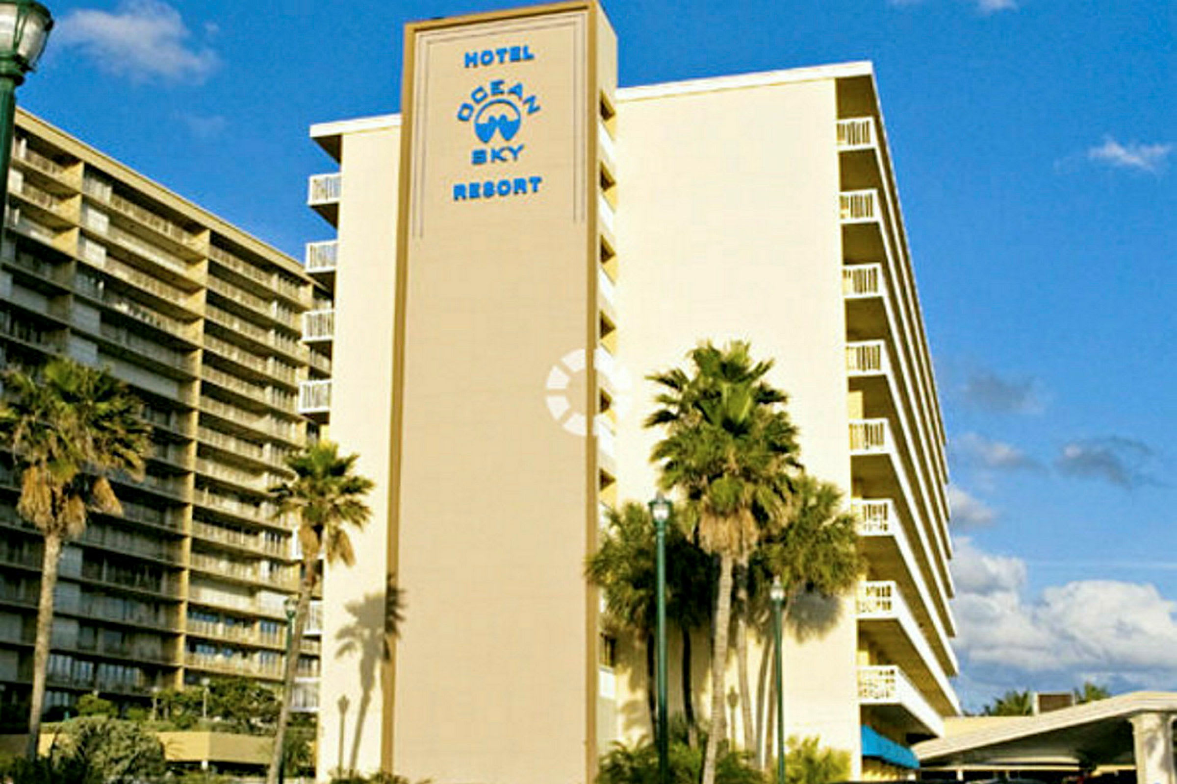 Et billede af et hotel med teksten "Hotel Ocean Sky Resort" på facaden. Der ses palmer foran bygningen. (AI-genereret tekst)