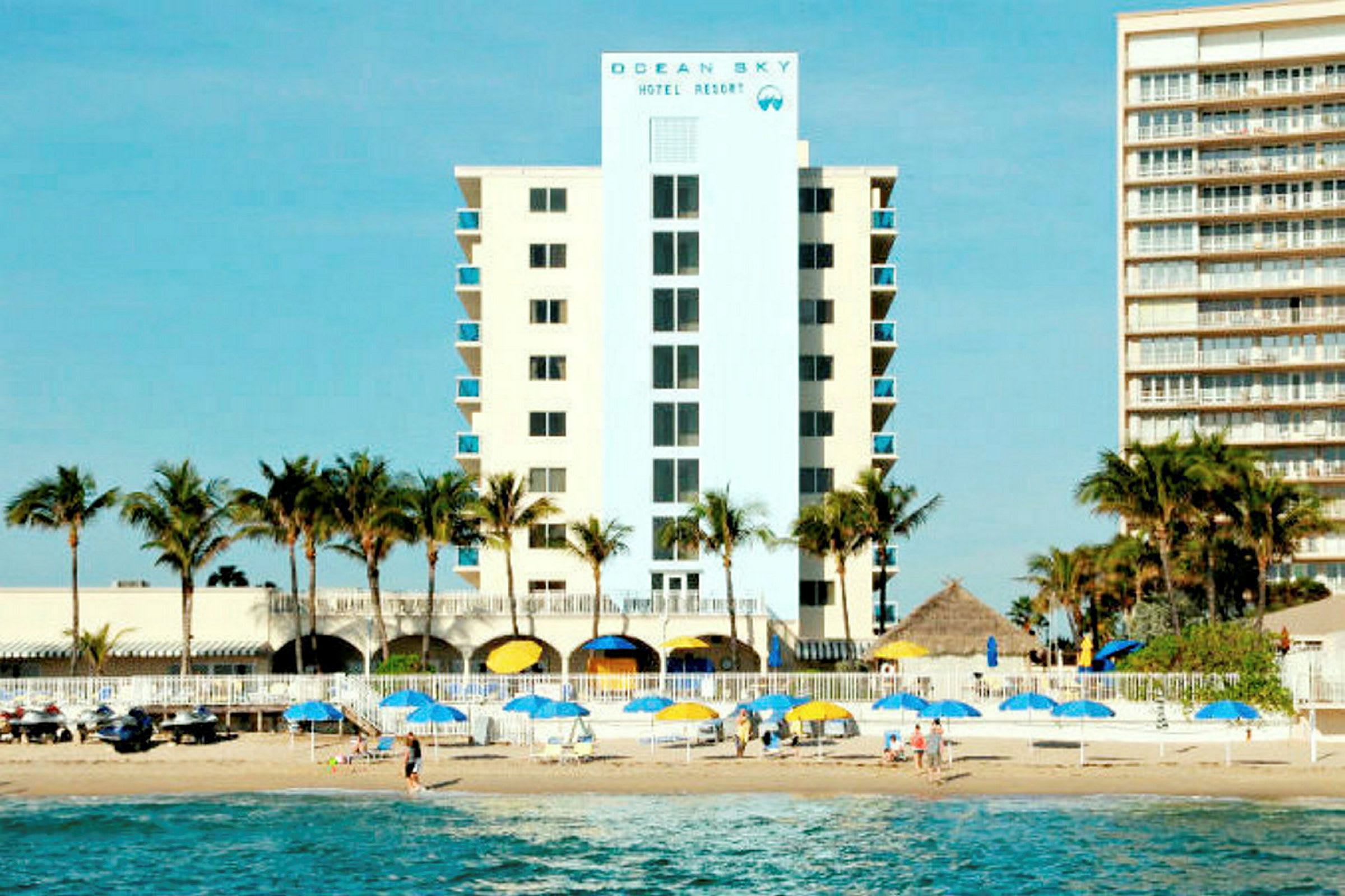 Et hotel med palmer foran, ved siden af stranden med parasoller og mennesker på sanden. (AI-genereret tekst)