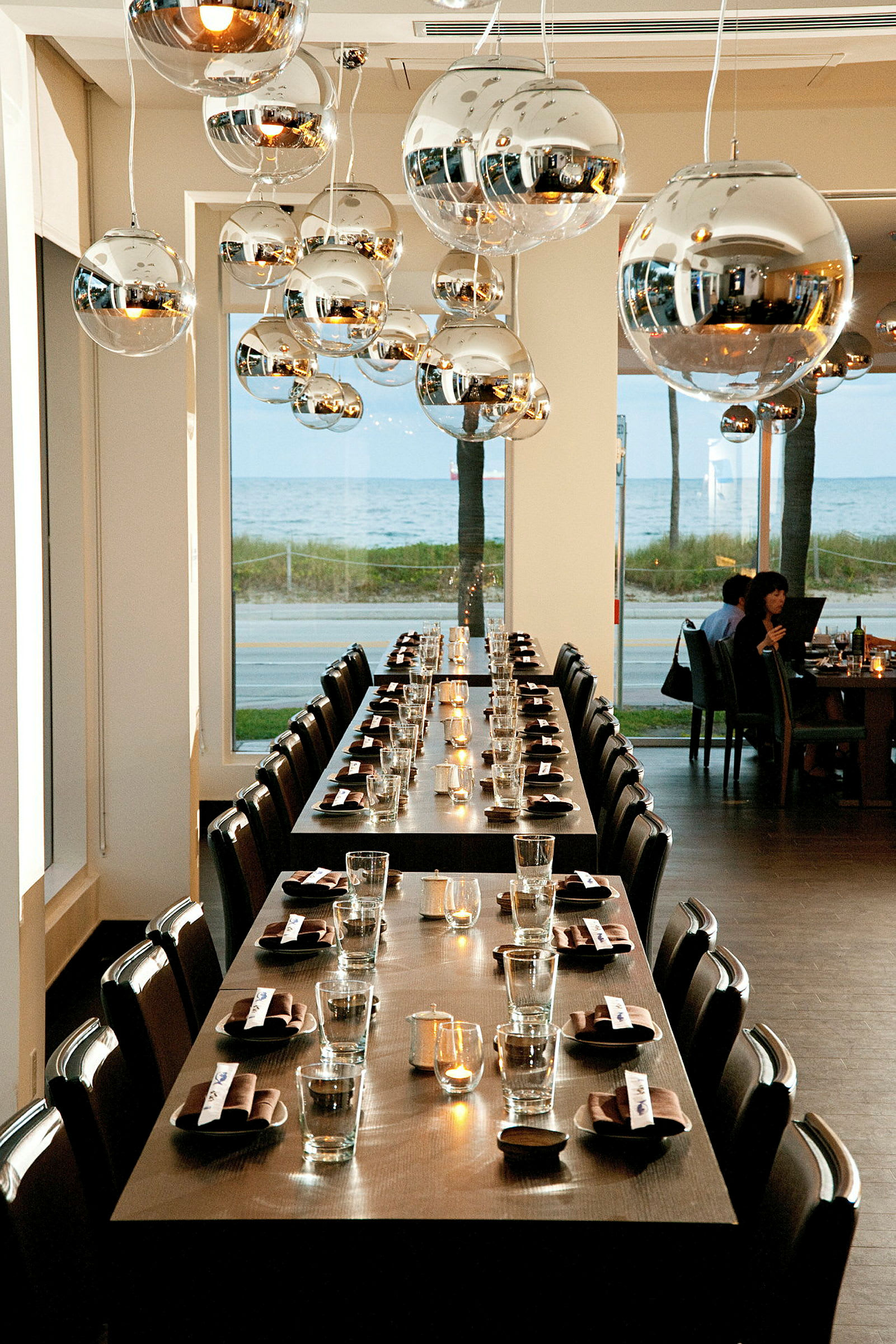 Billedet viser en restaurant med langborde, der er dækket med glas og servietter. Loftet har moderne metaliske lysekroner, og der er høje vinduer med udsigt over havet. (AI-genereret tekst)