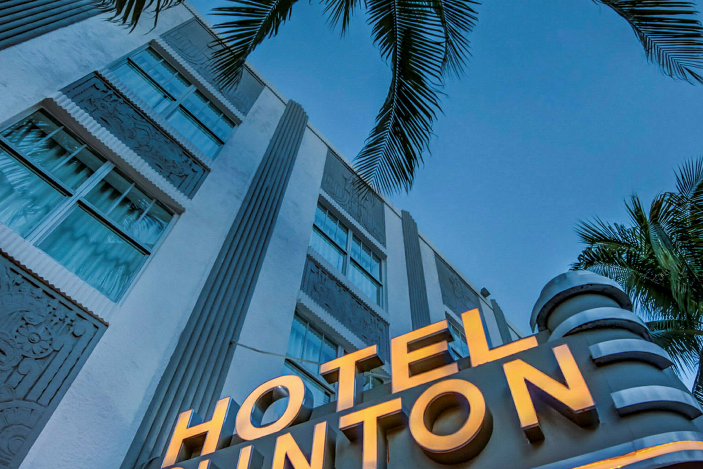 Et nærbillede af en hvid bygning med et oplyst skilt, der siger "HOTEL", og palmer i forgrunden mod en klar blå himmel. (AI-genereret tekst)
