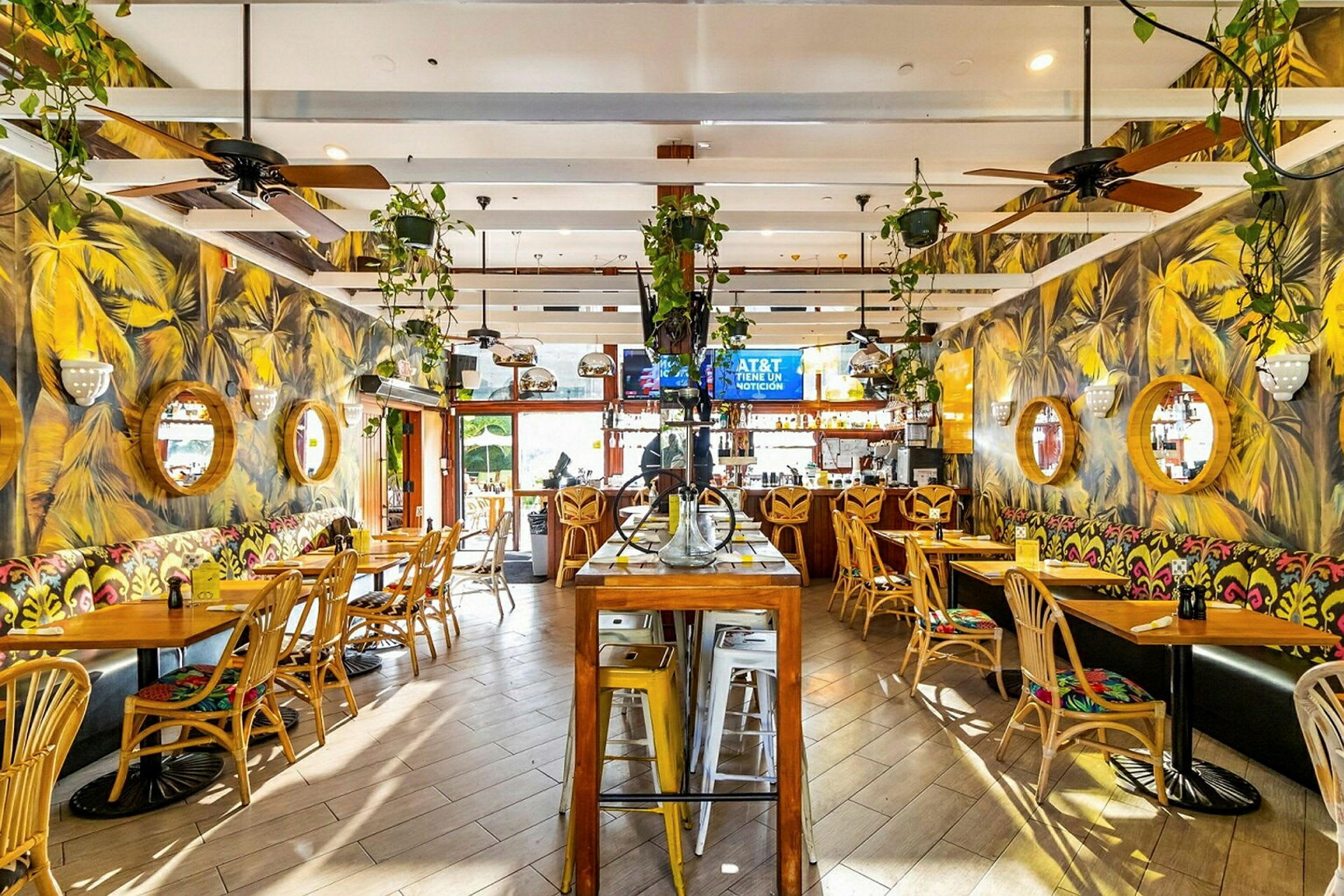 Indre af en restaurant med træstole, borde og farverige vægmalerier. Væggene er udsmykket med runde spejle, og planter hænger fra loftet. (AI-genereret tekst)