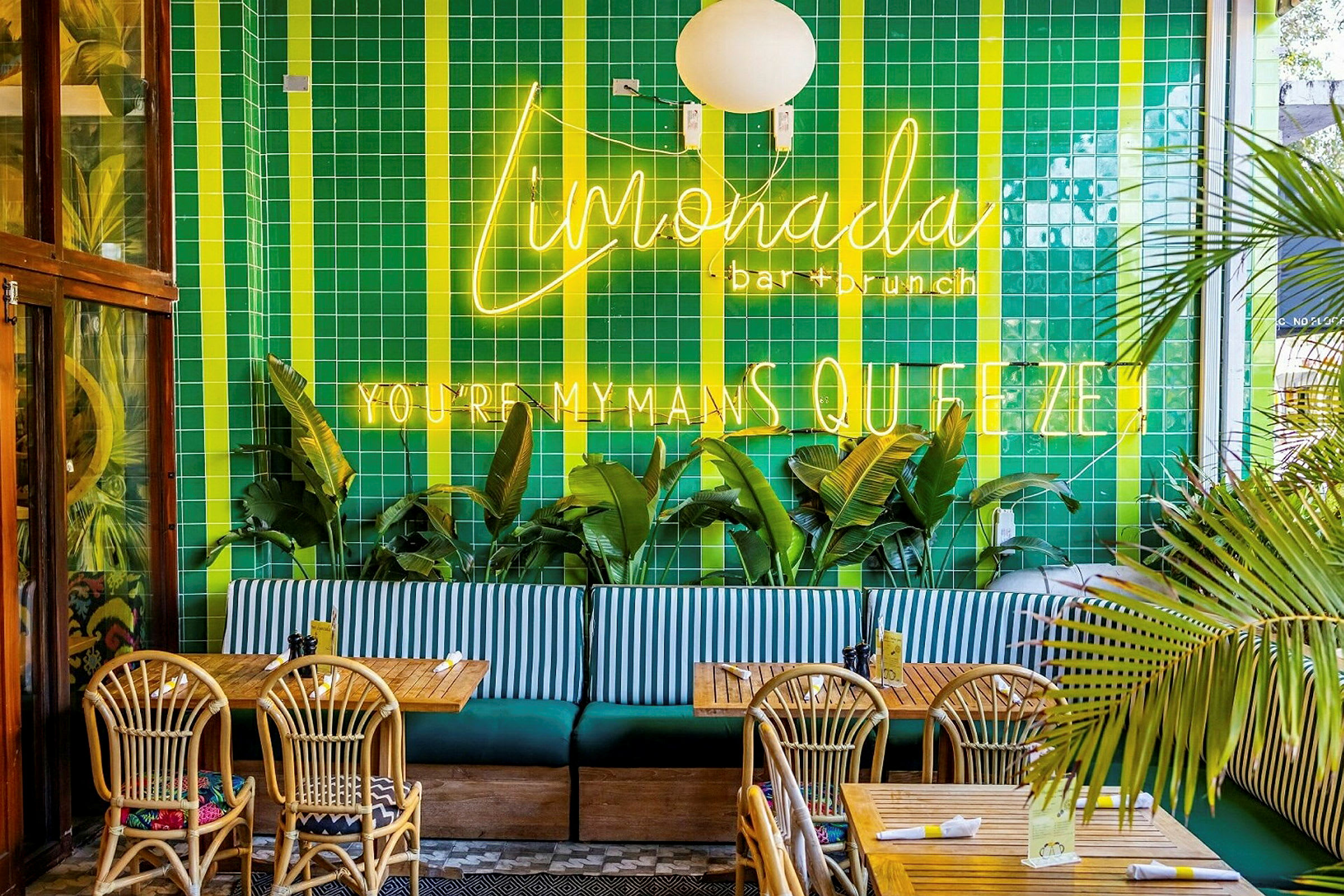 Restaurantinteriør med grønne flisebelagte vægge, neonskilte, palmer, træborde og flettede stole. (AI-genereret tekst)