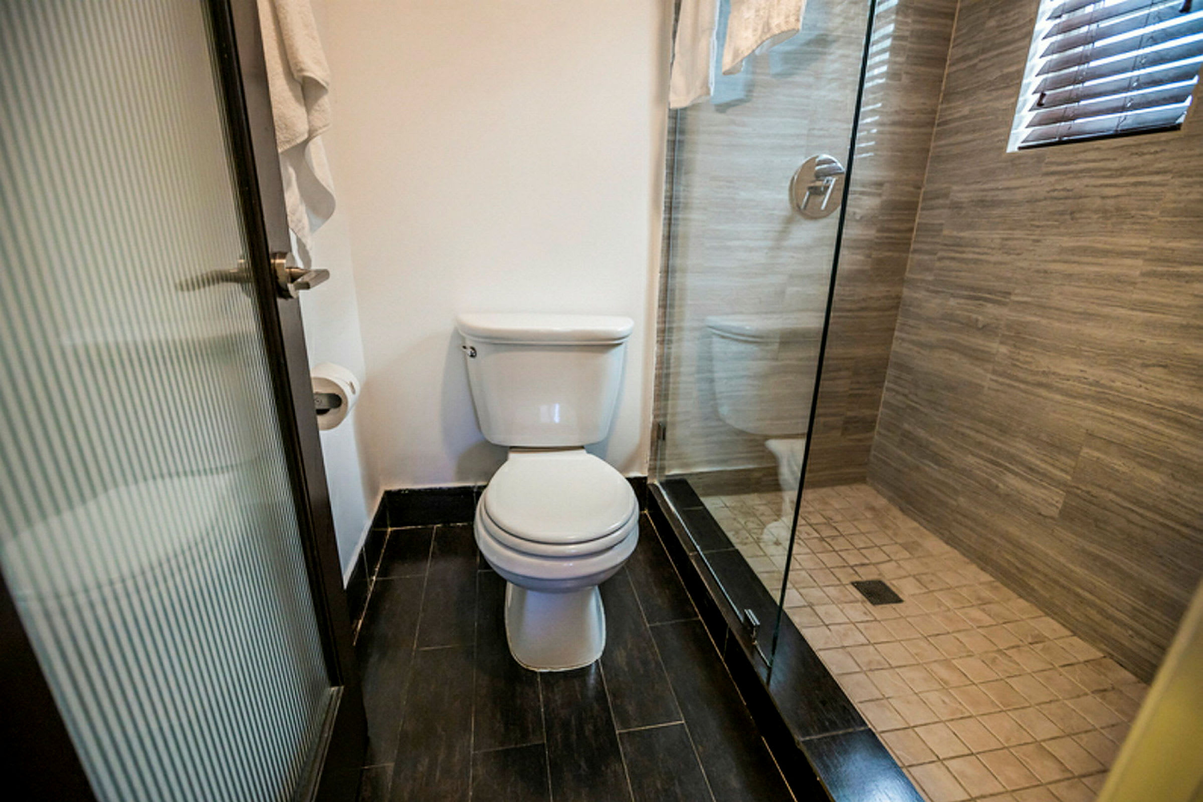 Et badeværelse med et toilet og en bruser, bruserkabinen har glasvægge og gulvet er dækket med mørke fliser. (AI-genereret tekst)