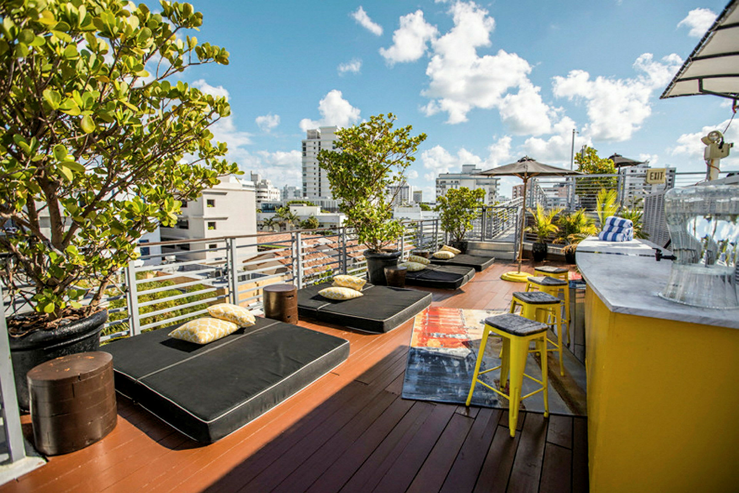 En terrasse med liggestole, puder og grønne planter. Der er en gul bardisk med taburetter ved siden af. (AI-genereret tekst)