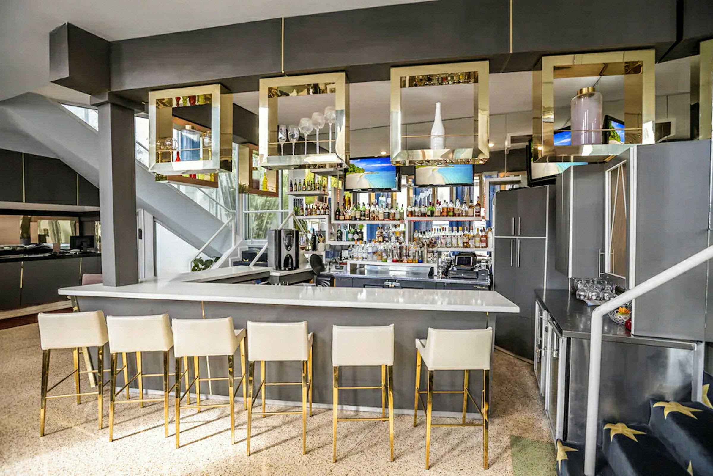 En moderne bar med hvid bardisk og høje stole i lys farve med gyldne ben. Over bardisken hænger firkantede dekorationer. (AI-genereret tekst)