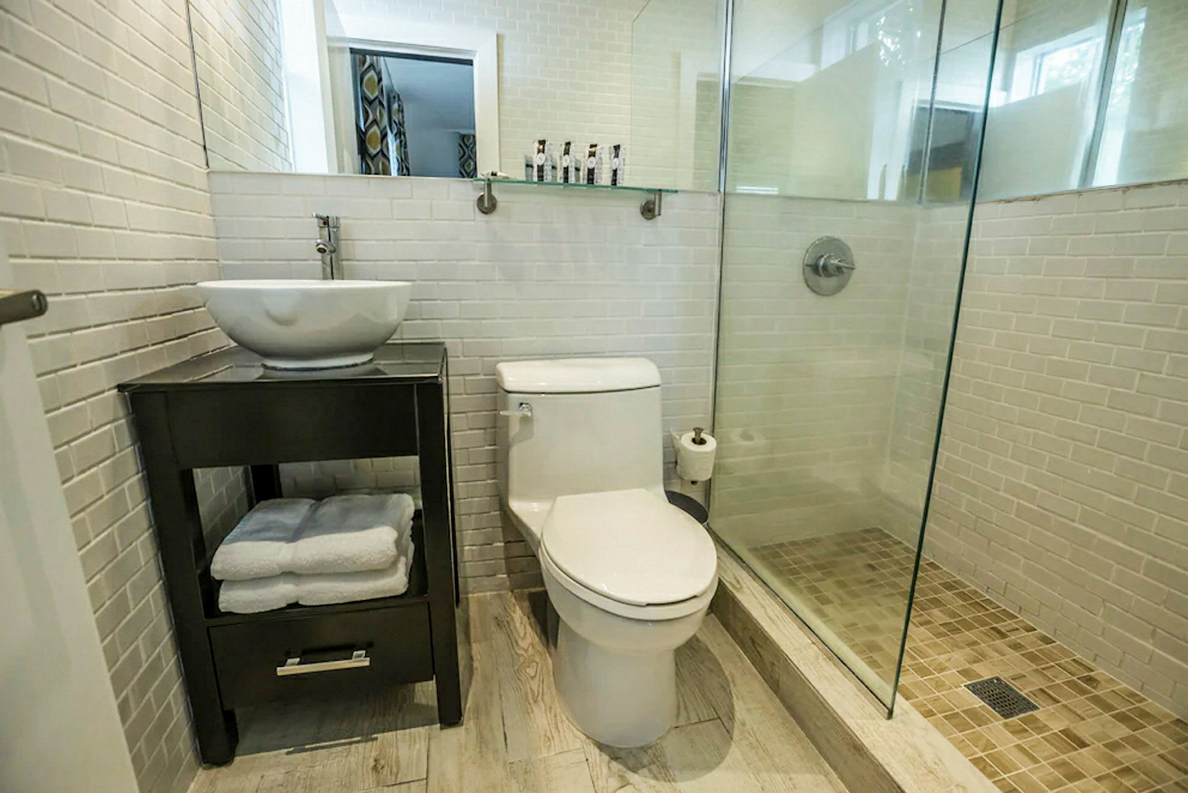 Et badeværelse med hvide fliser på væggene, en brusekabine med glasdør, et toilet og en håndvask på et mørkt skab. (AI-genereret tekst)