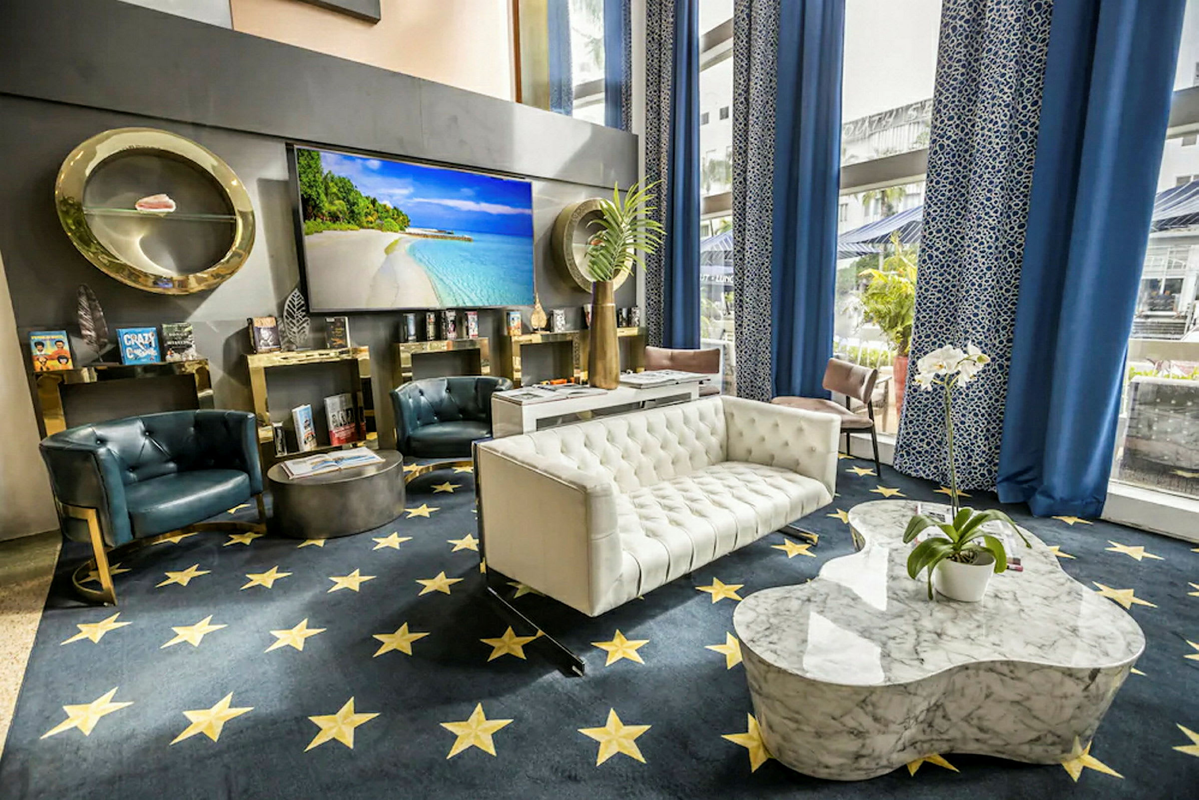 Billede af en elegant hotellobby med sofaer, lænestole og et marmorbord. I baggrunden ses en stor skærm med strandmotiv, og indretningen omfatter blå gardiner og dekorative detaljer. (AI-genereret tekst)