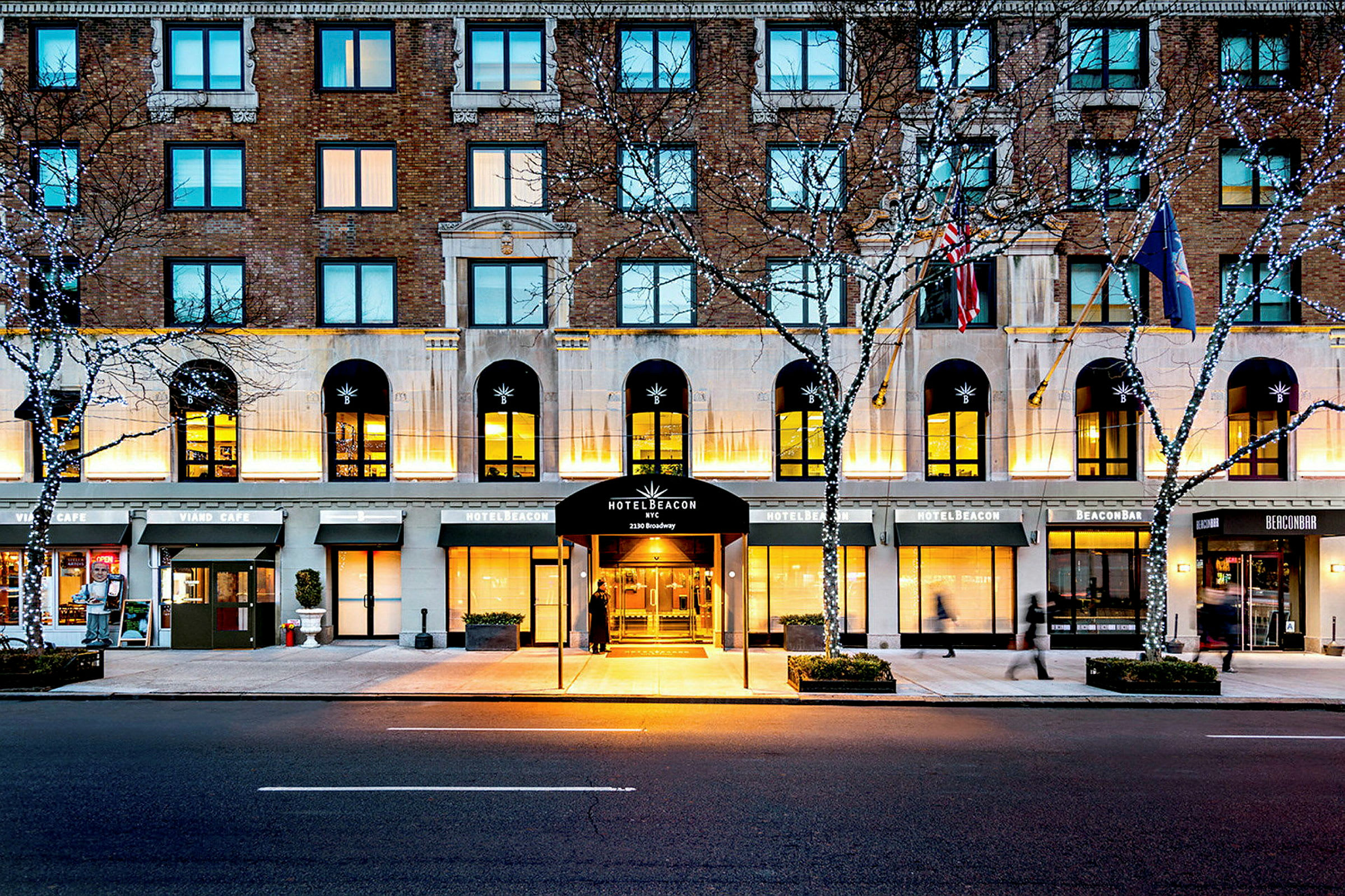 Hotelbygning ved en vej, med indgang markeret som Hotel Beacon, omgivet af cafémiljø og lyskæder i træerne. (AI-genereret tekst)
