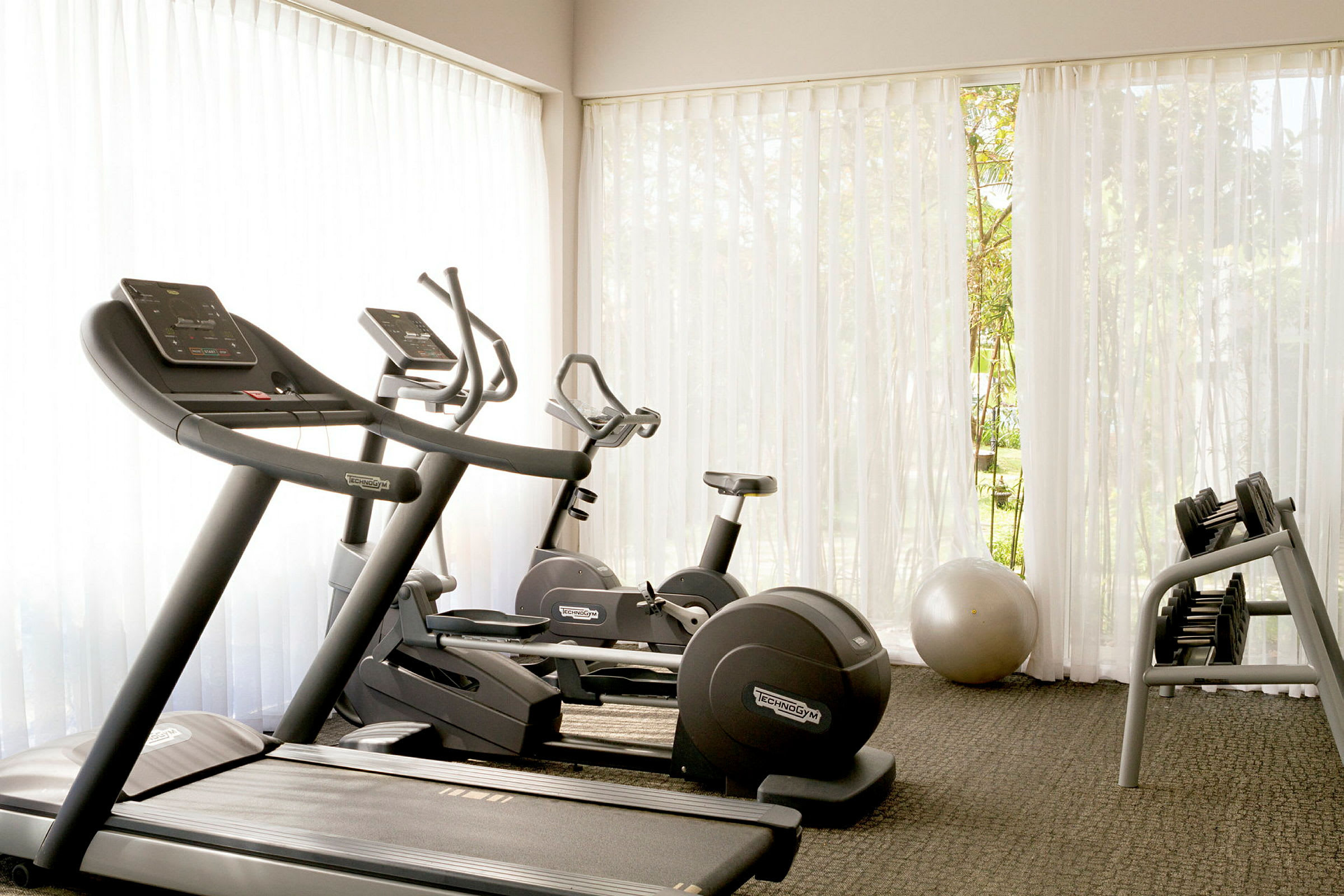 Et fitnesscenter med træningsmaskiner, inklusive løbebånd, motionscykel og håndvægte, med naturligt lys, der strømmer gennem store vinduer. Hvide gardiner skaber en lys og luftig atmosfære. (AI-genereret tekst)