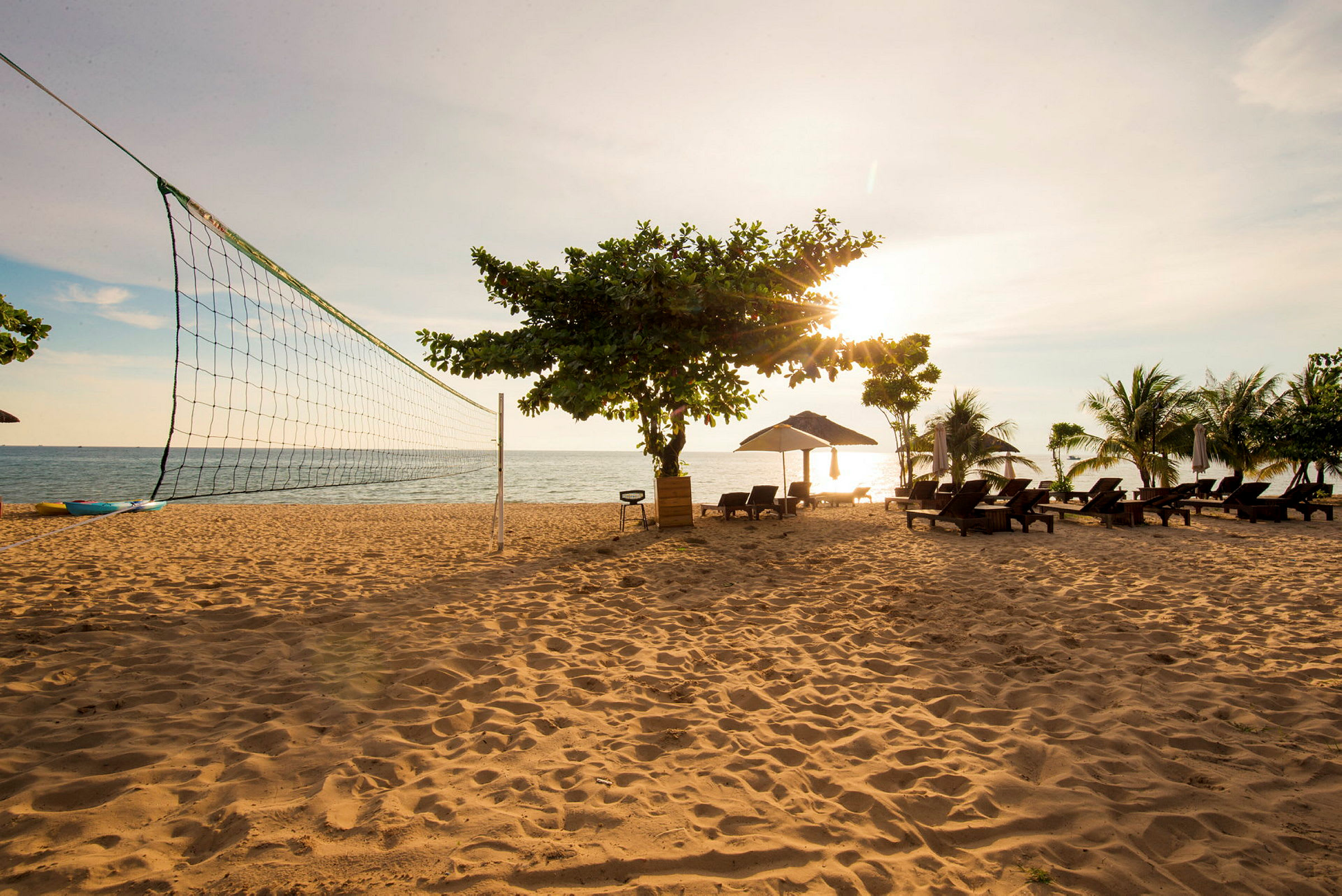 En sandstrand med et volleyballnet, solsenge under parasoller og nogle træer. Havet ses i baggrunden under en solnedgang. (AI-genereret tekst)