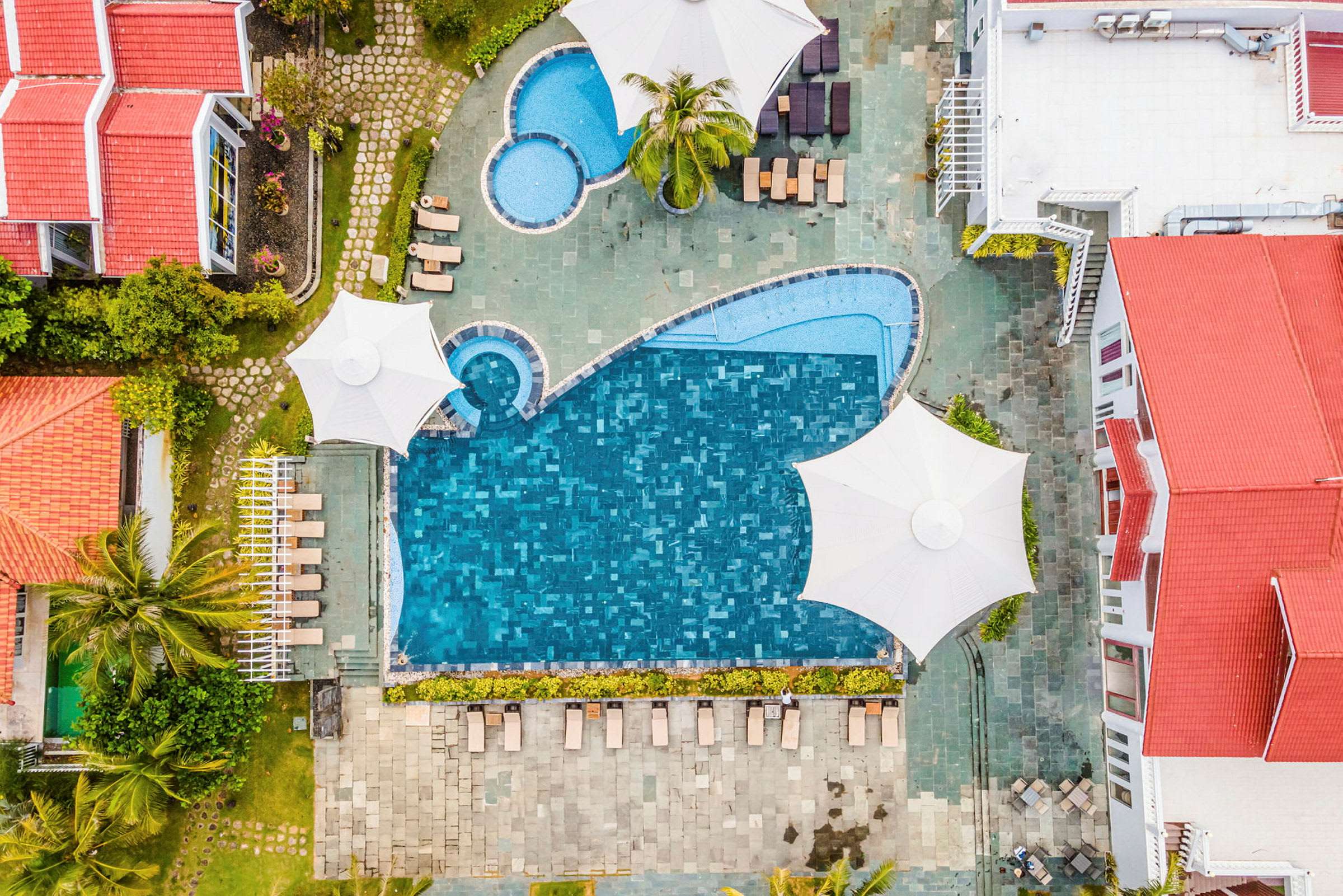 Luftfoto af en udendørs pool omgivet af liggestole, parasoller og bygninger med røde tage. (AI-genereret tekst)
