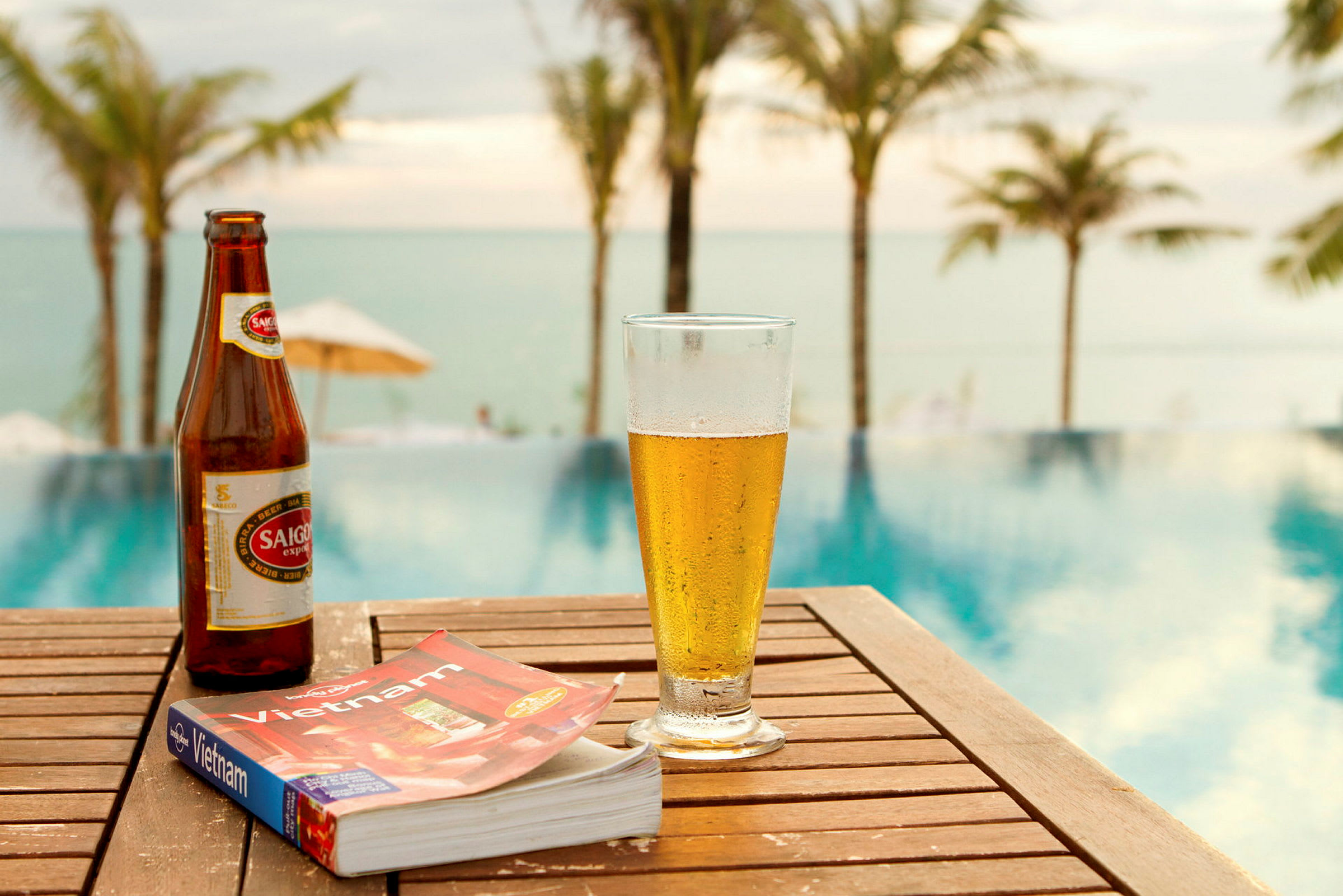 En flaske øl, et glas øl og en bog med titlen "Vietnam" står på et træbord ved en pool. I baggrunden ses palmer og hav. (AI-genereret tekst)