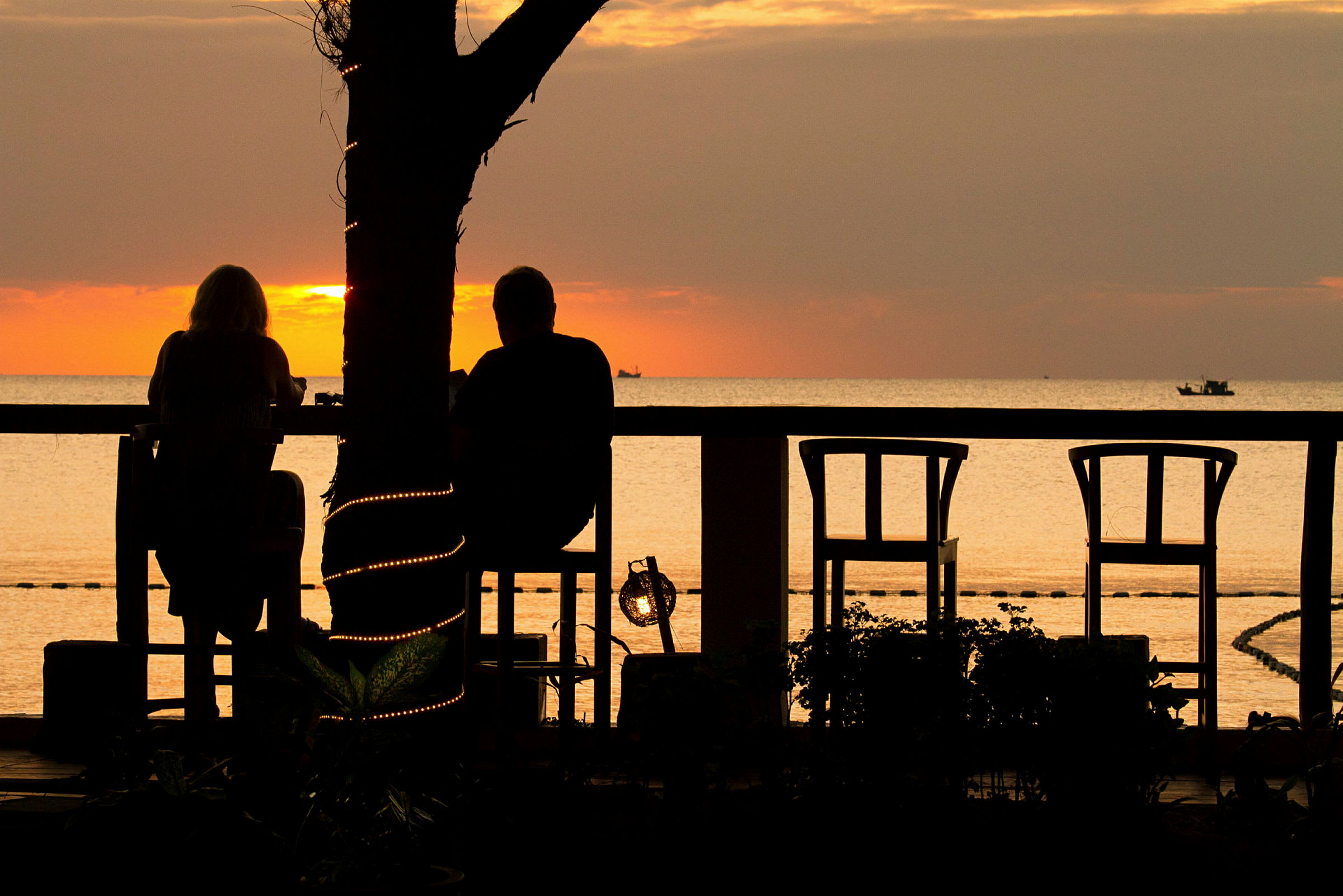 To personer sidder ved et bord og nyder solnedgangen ved havet. Et træ med dekorative lys står i forgrunden. (AI-genereret tekst)