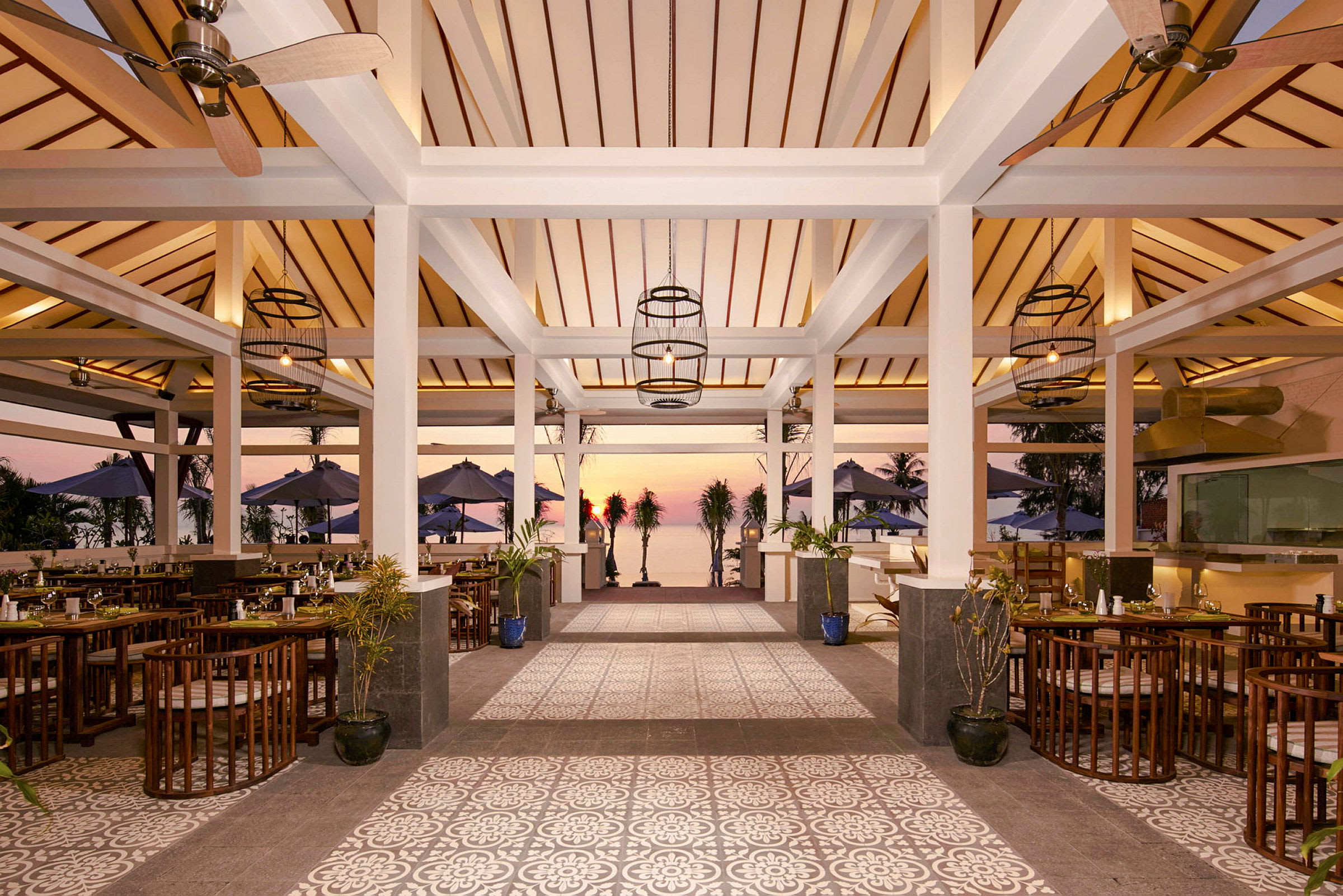 En restaurant med trætag, åbne vægge og gulv med dekorative mønstre. Udenfor ses borde, stole og parasoller samt solnedgang ved havet. (AI-genereret tekst)