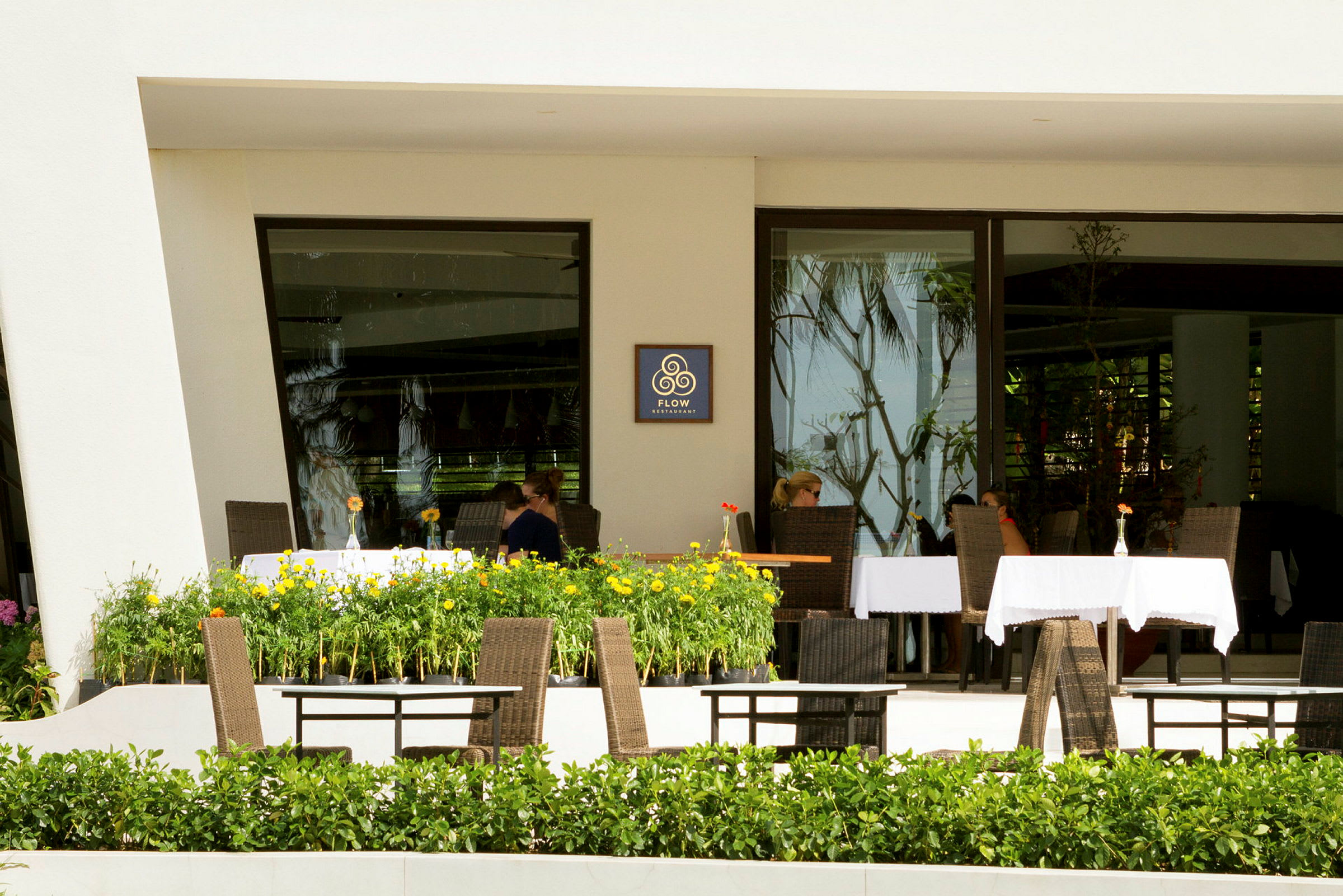 En restaurant med borde og stole på en terrasse omgivet af grønne planter og blomster. (AI-genereret tekst)
