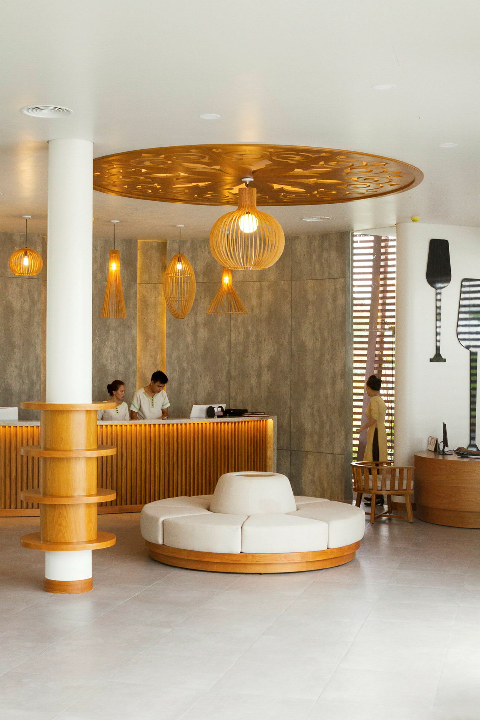 En hotellreception med moderne og stilfuldt interiør. Der er en skranke med personale, en rund siddeplads i midten af rummet og dekorative lamper i loftet. (AI-genereret tekst)