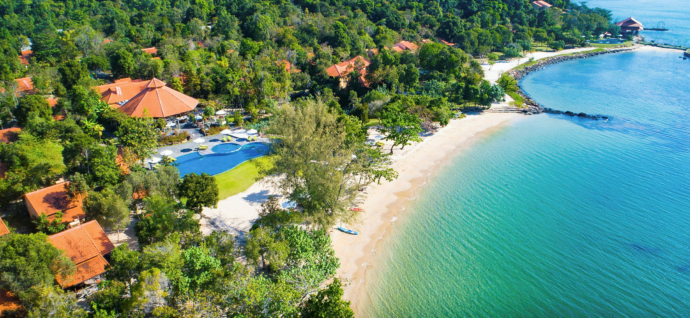 Luftfoto af strand med klart vand, omgivet af frodige træer og bygninger med røde tage. (AI-genereret tekst)