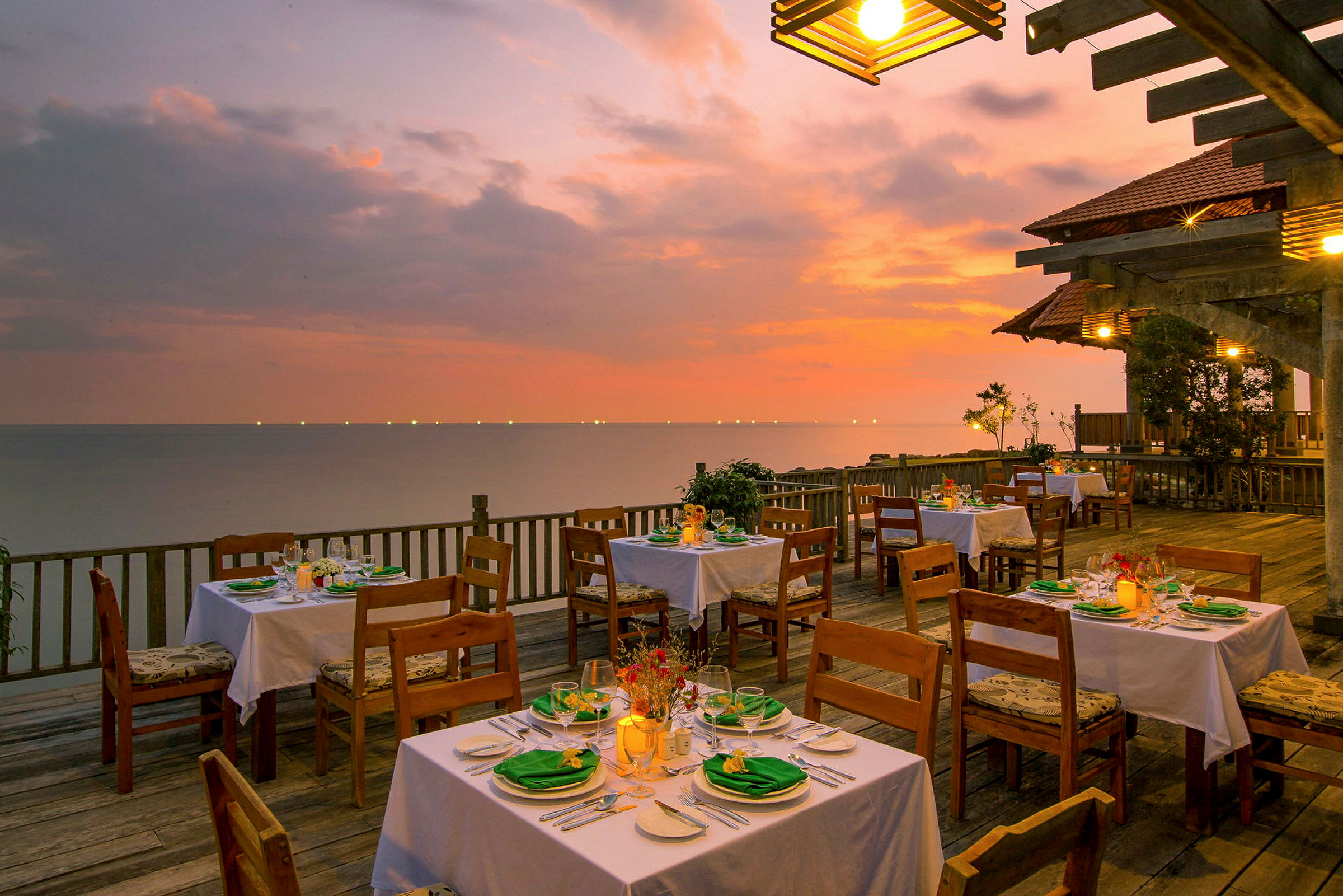 En udendørs restaurant med dækkede borde på en træterrasse ved havet under en farverig solnedgang. (AI-genereret tekst)