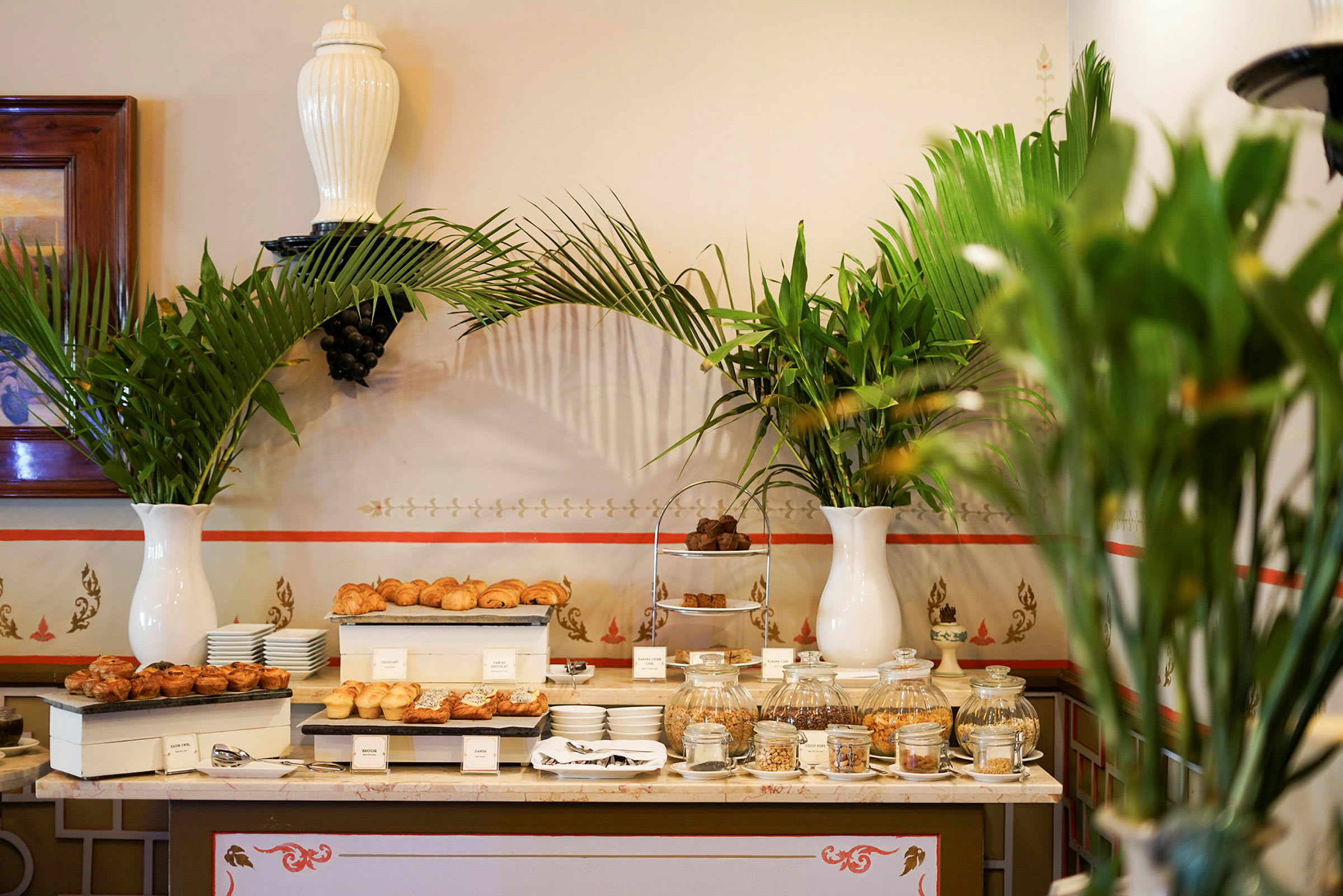 Billede af et bord med en buffet af bagværk og snacks, dekoreret med grønne planter i hvide vaser. (AI-genereret tekst)
