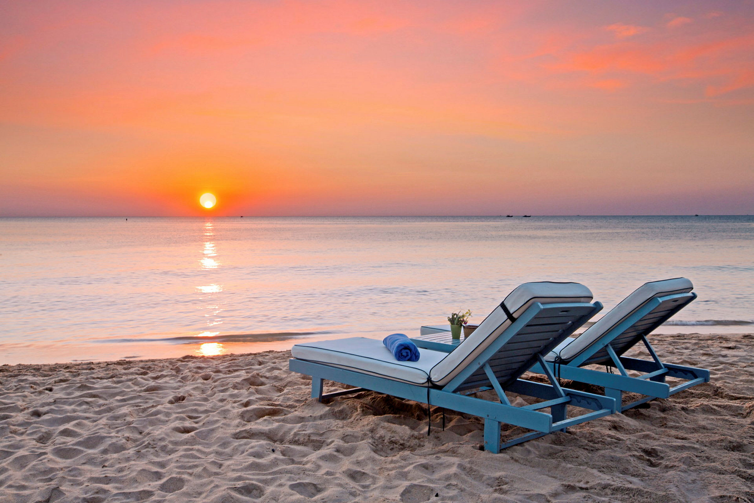 To liggestole står på sandstranden ved havet med en solnedgang i baggrunden. (AI-genereret tekst)