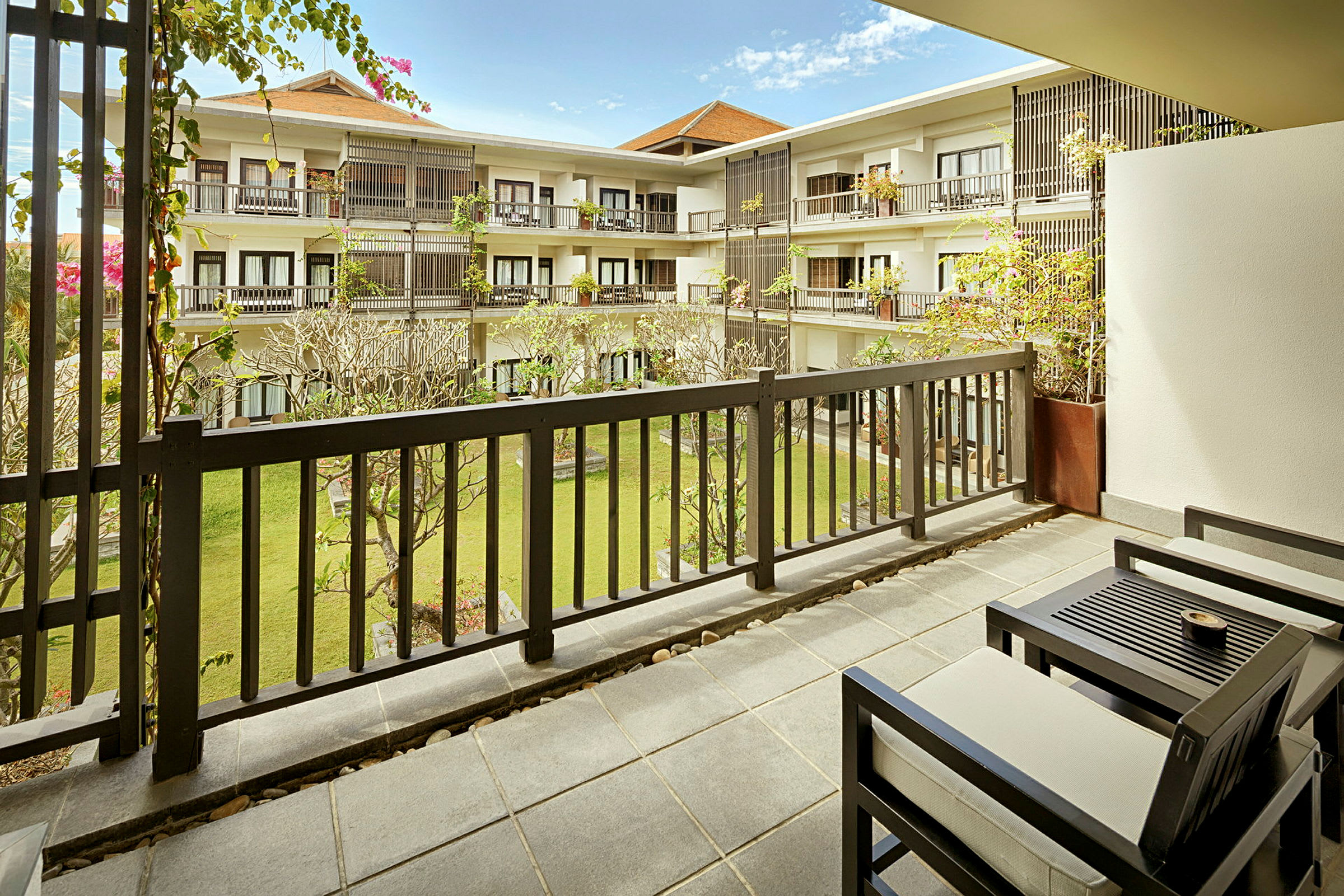 Udsigt fra en balkon med bord og stole mod en gårdhave med grønne områder og omkringliggende bygninger. (AI-genereret tekst)