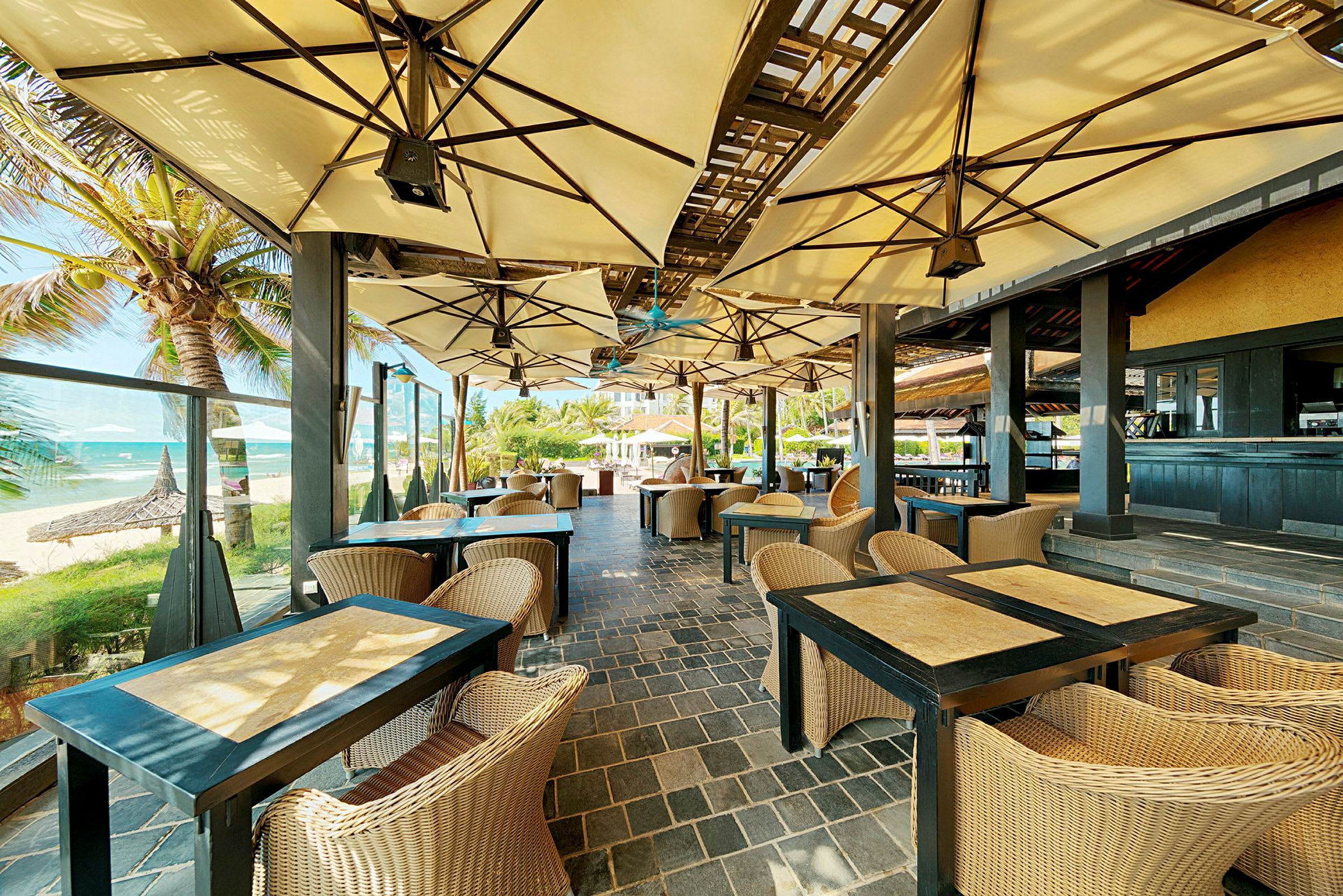 En restaurant med udendørs servering ved stranden med borde og stole under store parasoller. (AI-genereret tekst)