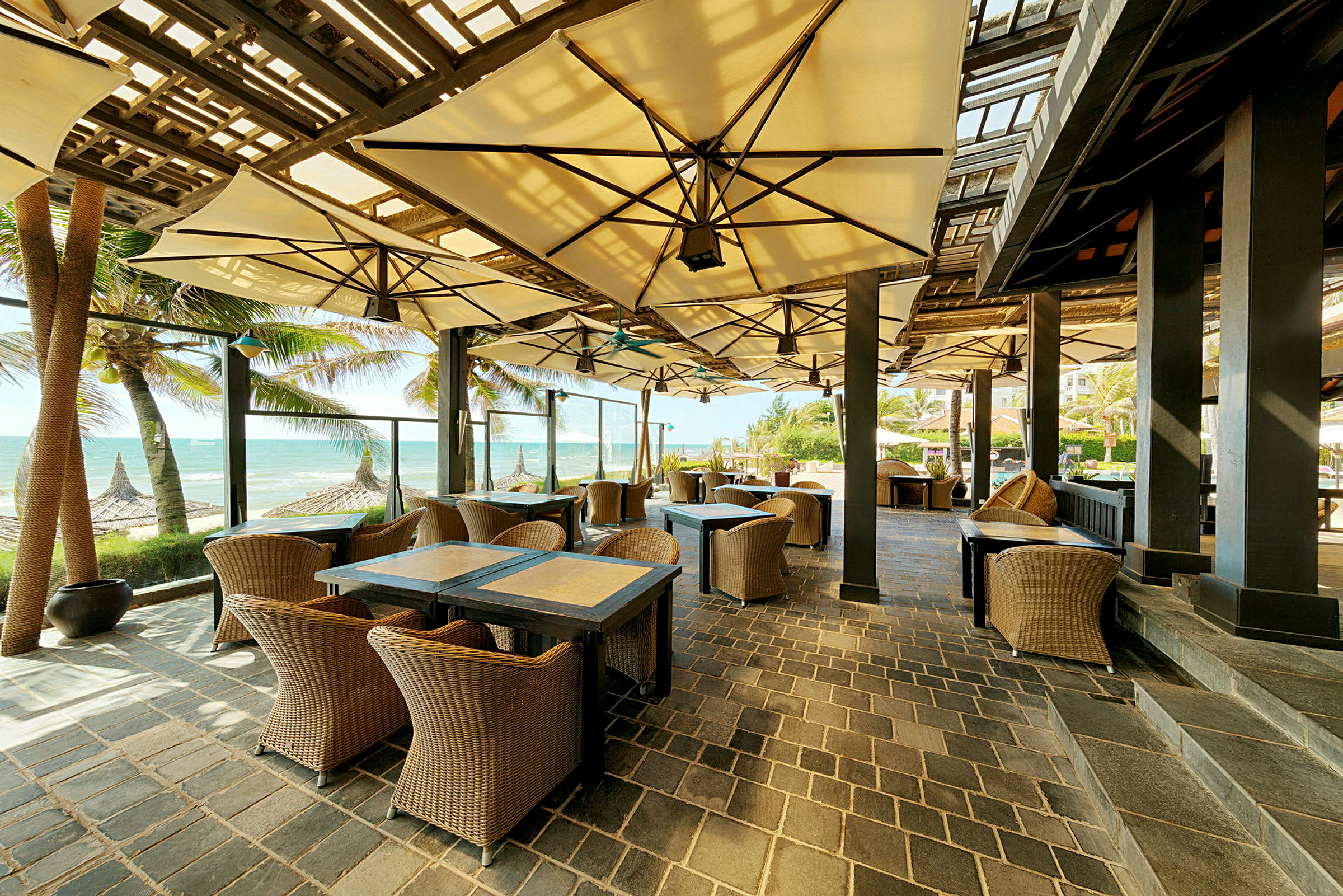 En udendørs restaurant med kurvemøbler og parasoller, beliggende ved stranden med udsigt over havet. (AI-genereret tekst)