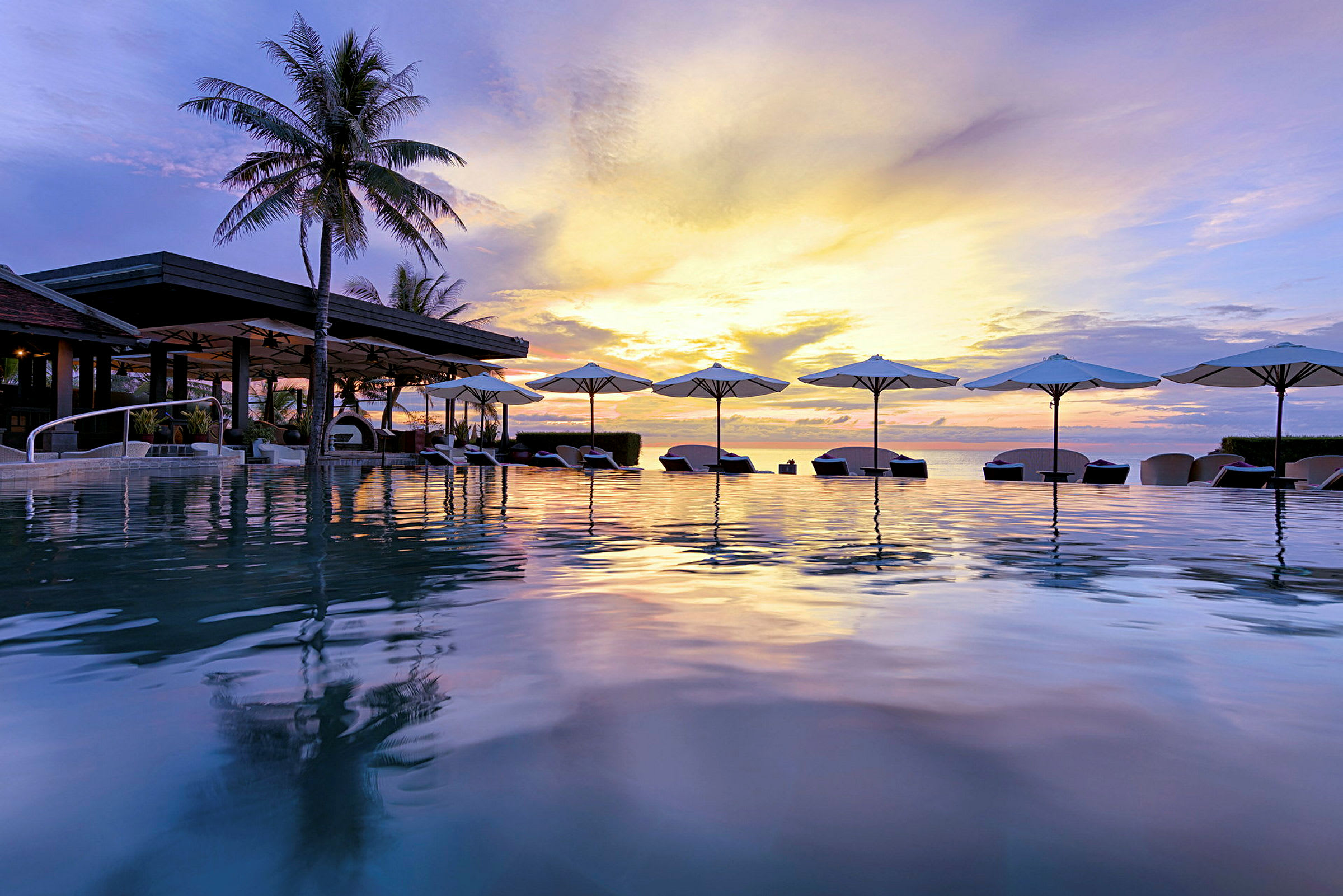 En infinitypool med liggestole og parasoller ved en tropisk resort, med en solnedgang der oplyser himlen. (AI-genereret tekst)