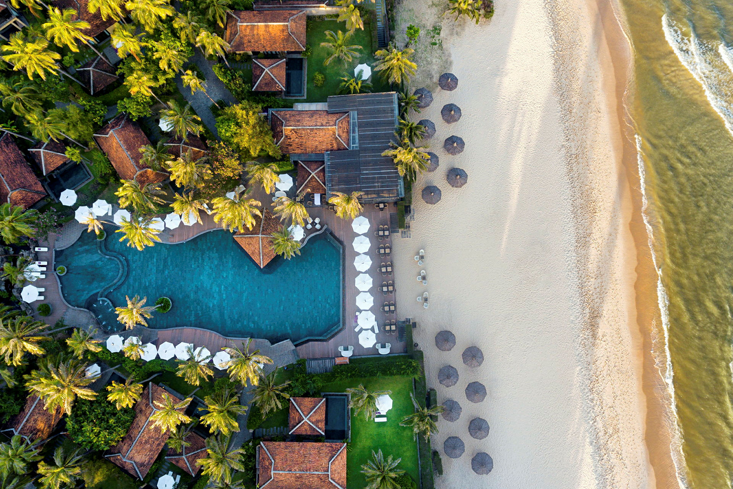 Luftfoto af et hotelområde med en pool, liggestole med parasoller og en sandstrand ved havet. (AI-genereret tekst)