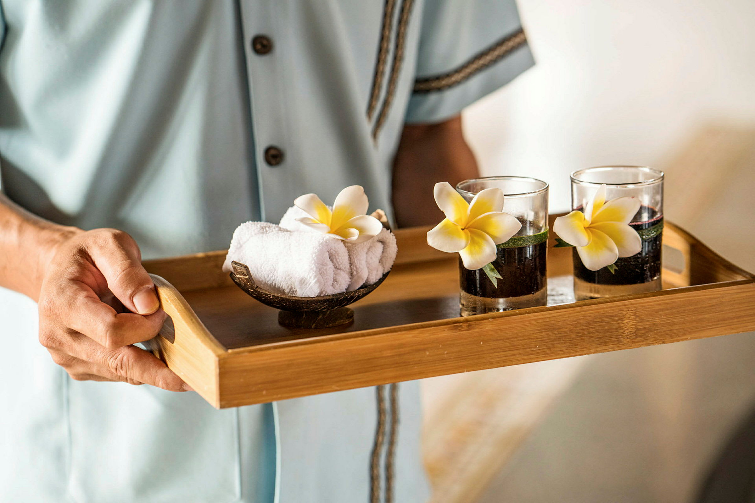 En person holder en træbakke med to glas dekoreret med blomster og en skål med rullede håndklæder. (AI-genereret tekst)