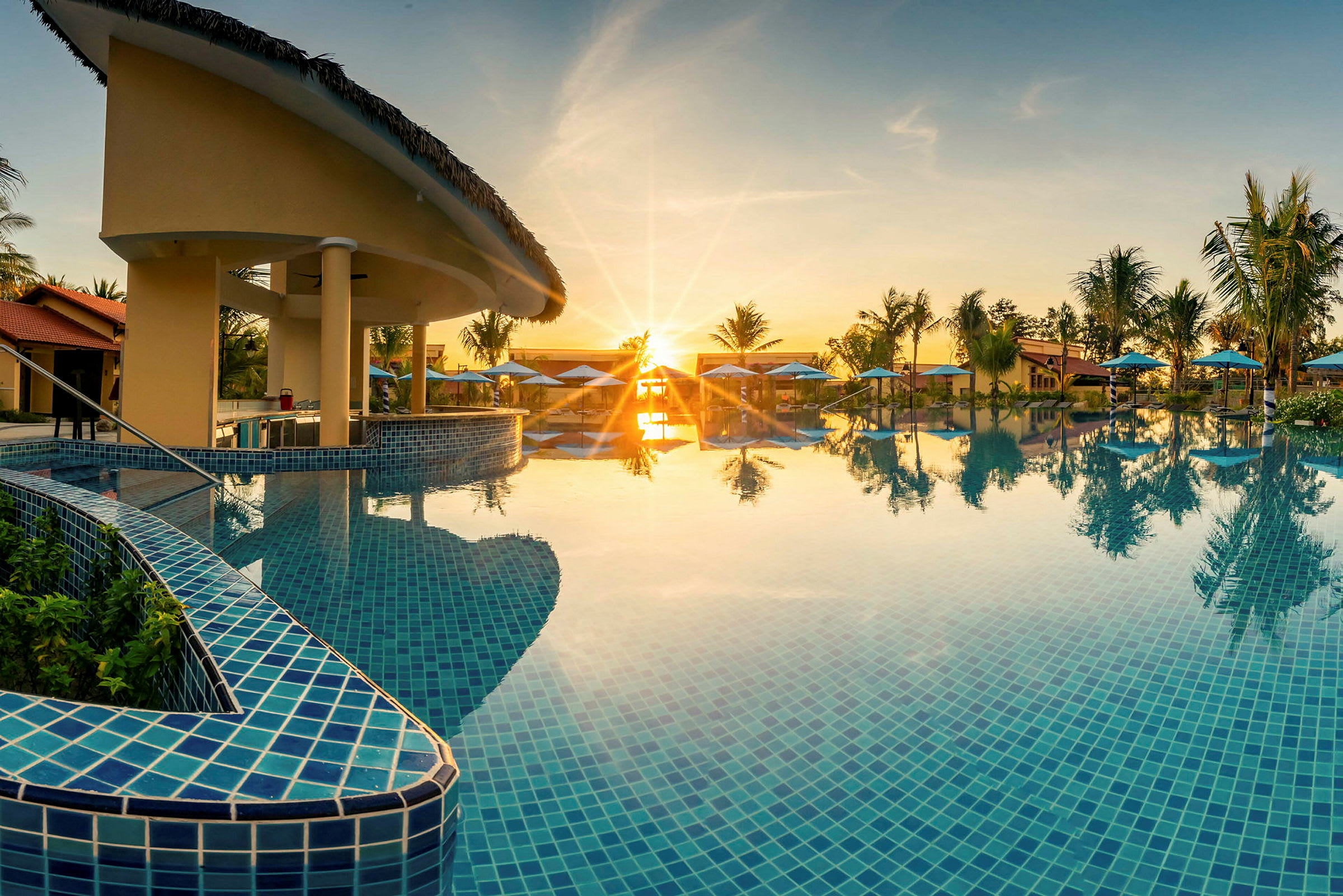En swimmingpool med blå fliser omgivet af palmer og parasoller ved solnedgang. (AI-genereret tekst)