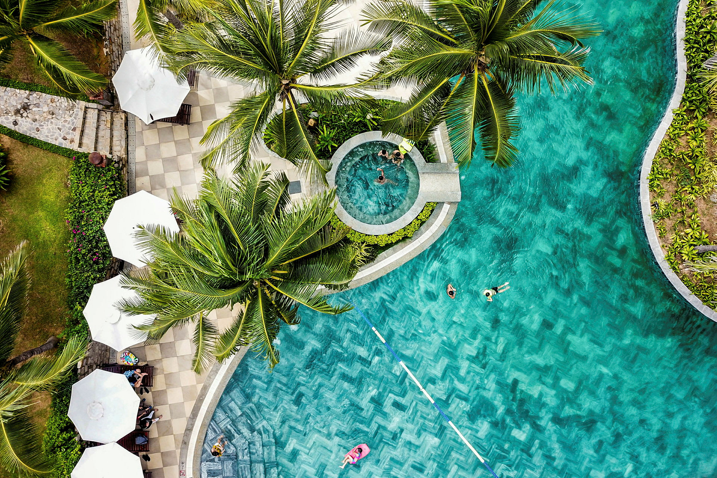 Et luftfoto af en pool omgivet af palmer og parasoller. Folk bader i poolen. (AI-genereret tekst)