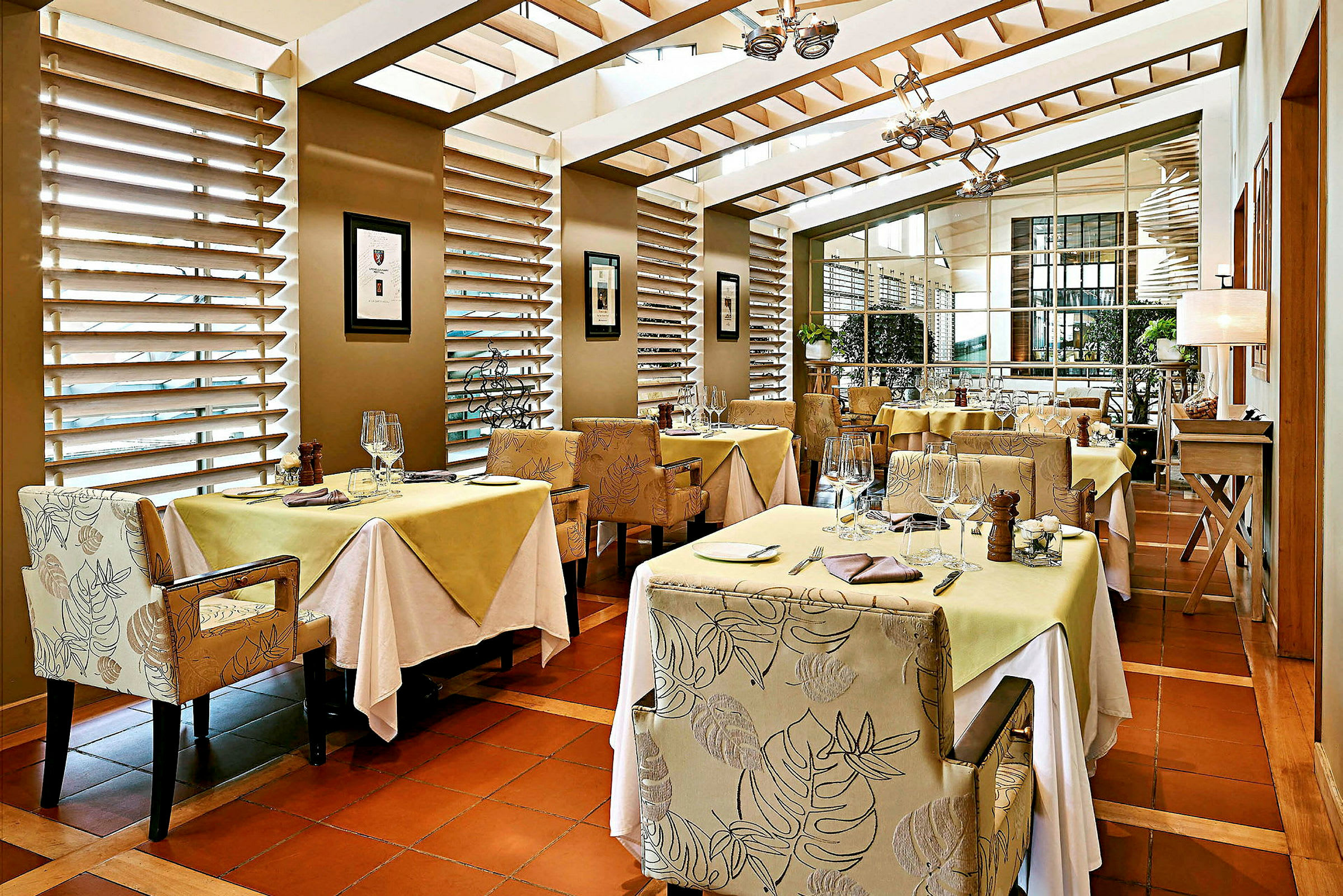 Et elegant restaurantområde med borde dækket til måltider, omgivet af stole med mønstret stof. Store vinduer lader naturligt lys komme ind. (AI-genereret tekst)