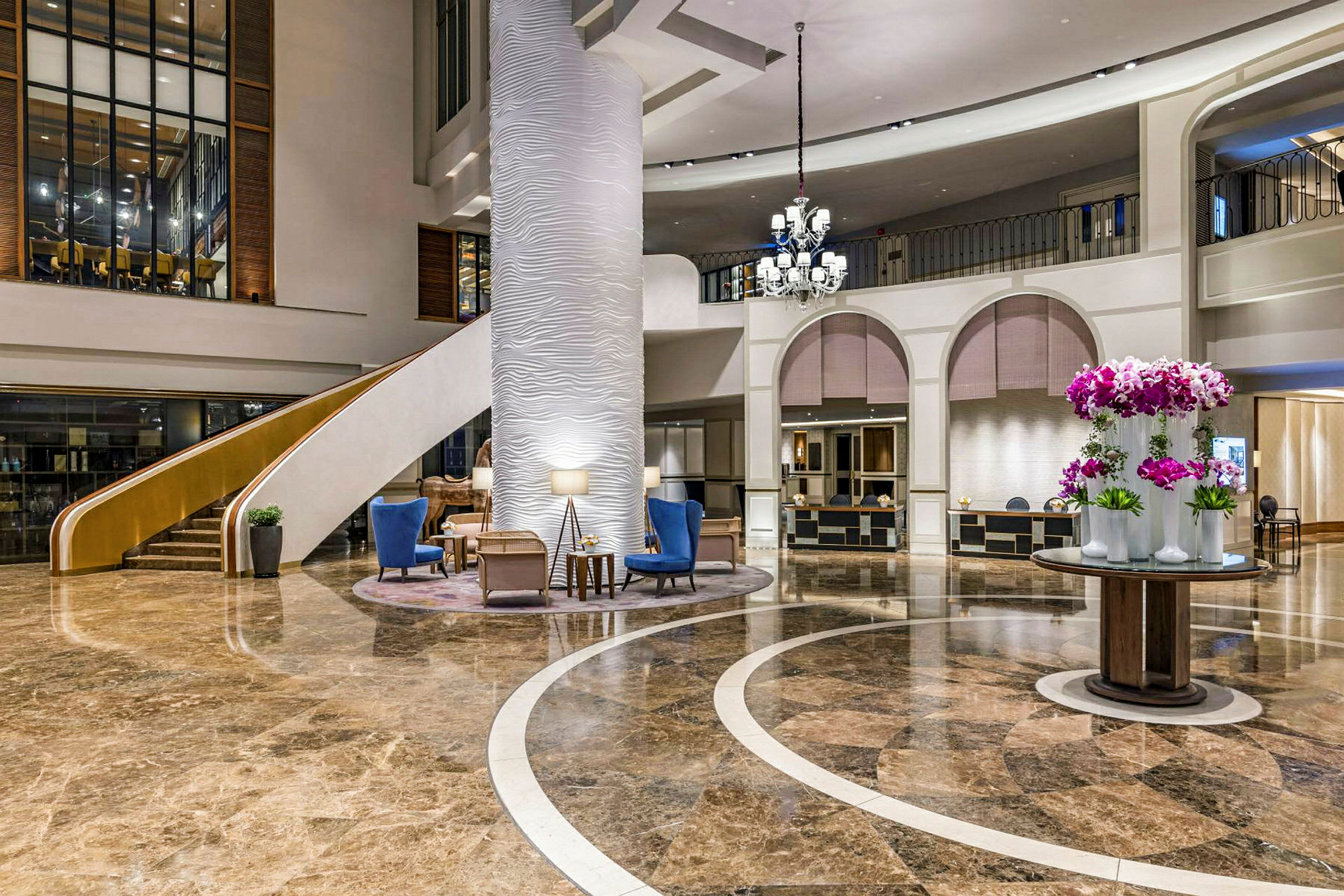 En hotellobby med marmorgulv, en stor søjle, en trappe, siddepladser og en rund borddekoration med blomster. (AI-genereret tekst)