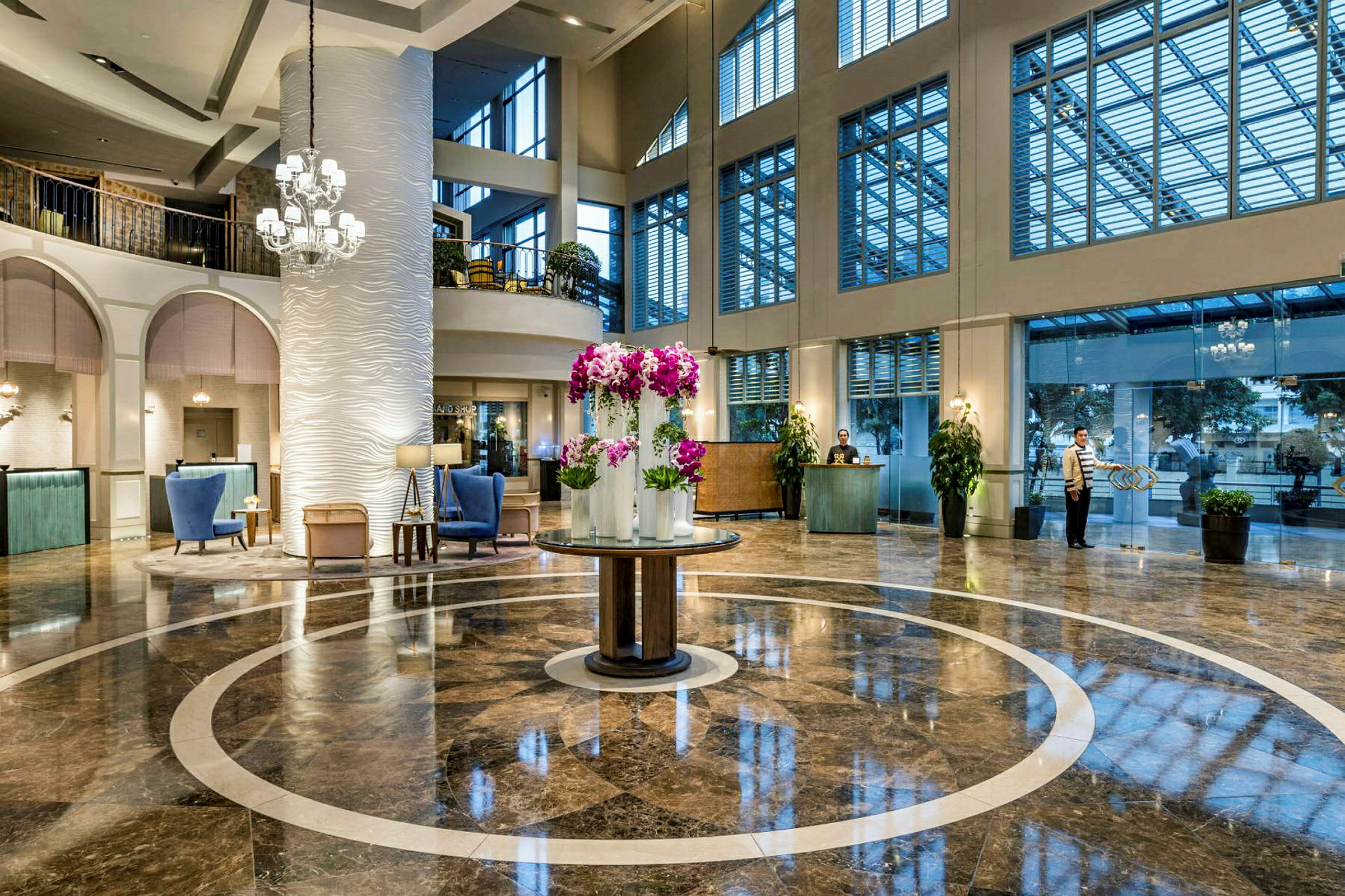 En elegant hotellobby med skinnende marmorgulv og et centralt rundt område dekoreret med lilla blomster i vaser. Høje vinduer slipper lys ind, og en reception ses i baggrunden. (AI-genereret tekst)