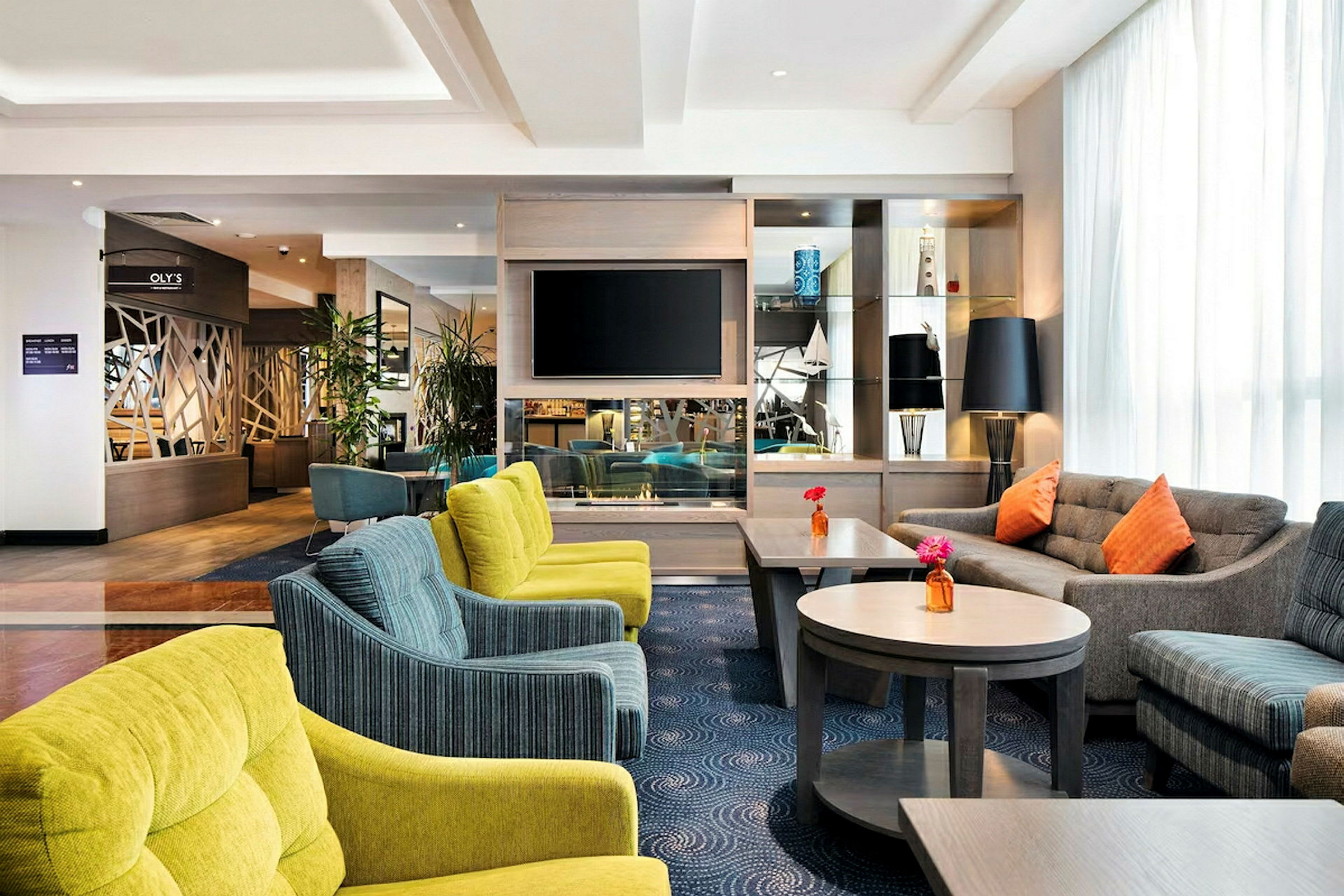 En hotellobby med komfortable siddeområder, flere sofaer og stole i forskellige farver, samt borde med blomster. Et TV er monteret på væggen, og der er dekorative genstande på hylderne. (AI-genereret tekst)