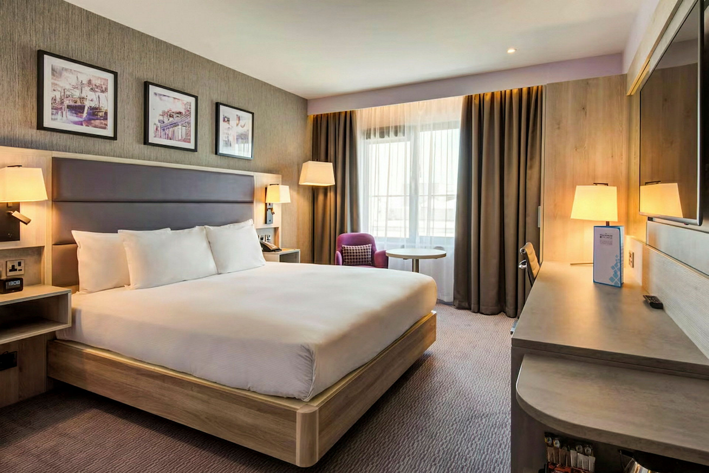 Hotelværelse med en stor seng, skrivebord, stol og vindue med gardiner. (AI-genereret tekst)