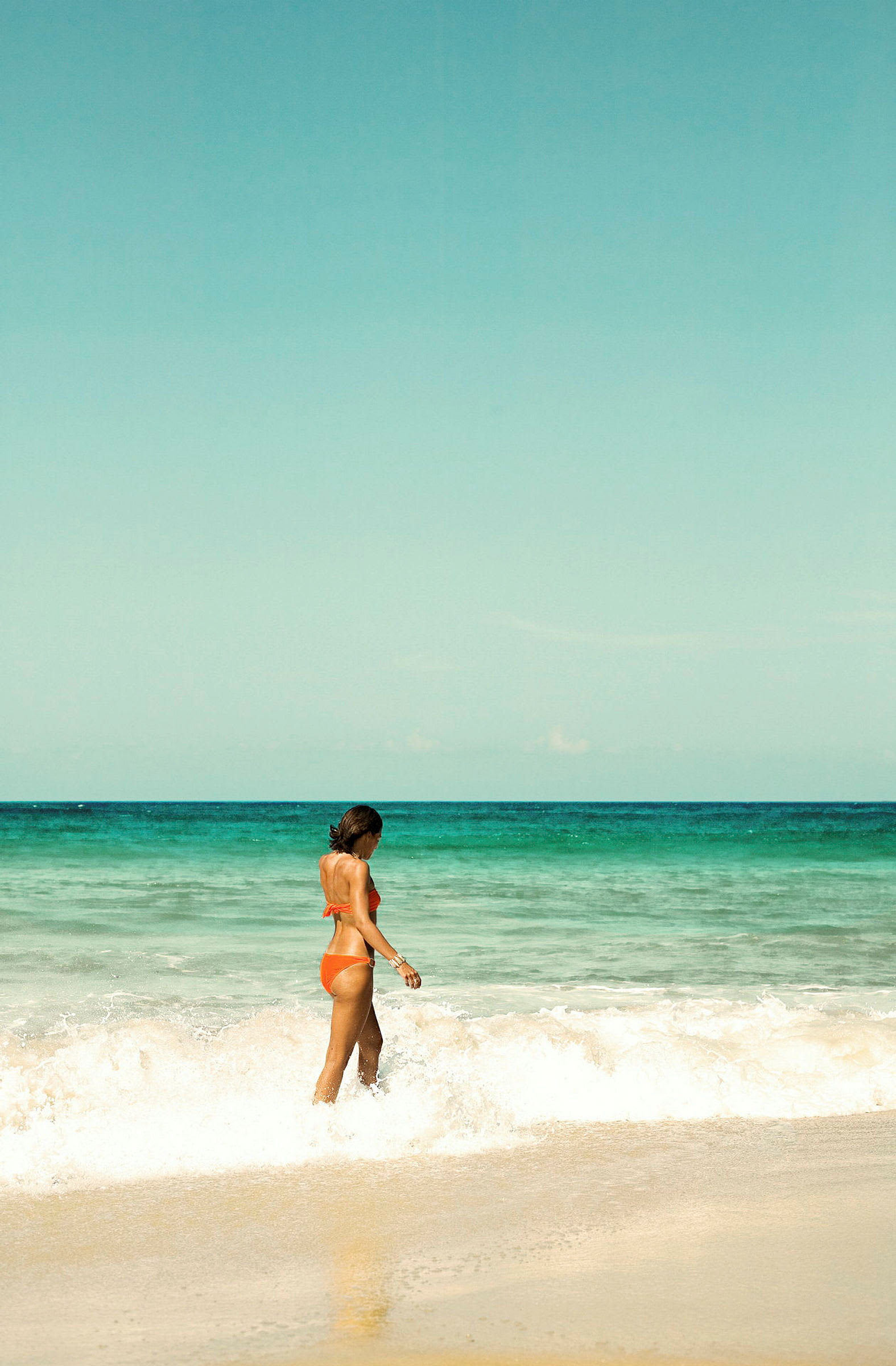En kvinde i orange bikini går ved stranden tæt på bølgerne. (AI-genereret tekst)