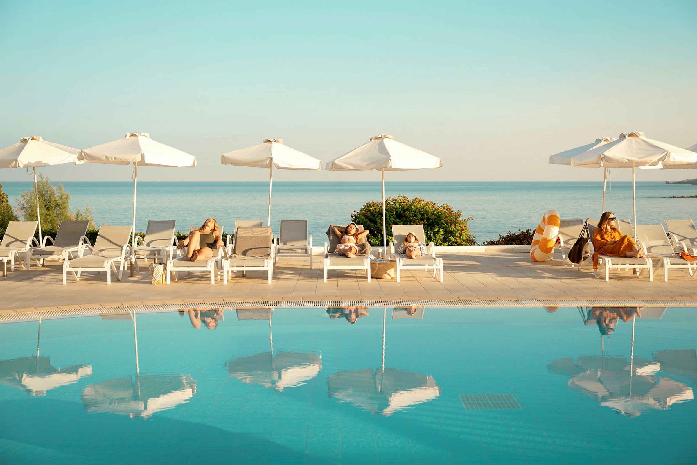 Personer slapper af på liggestole under parasoller ved en pool med havet i baggrunden. (AI-genereret tekst)