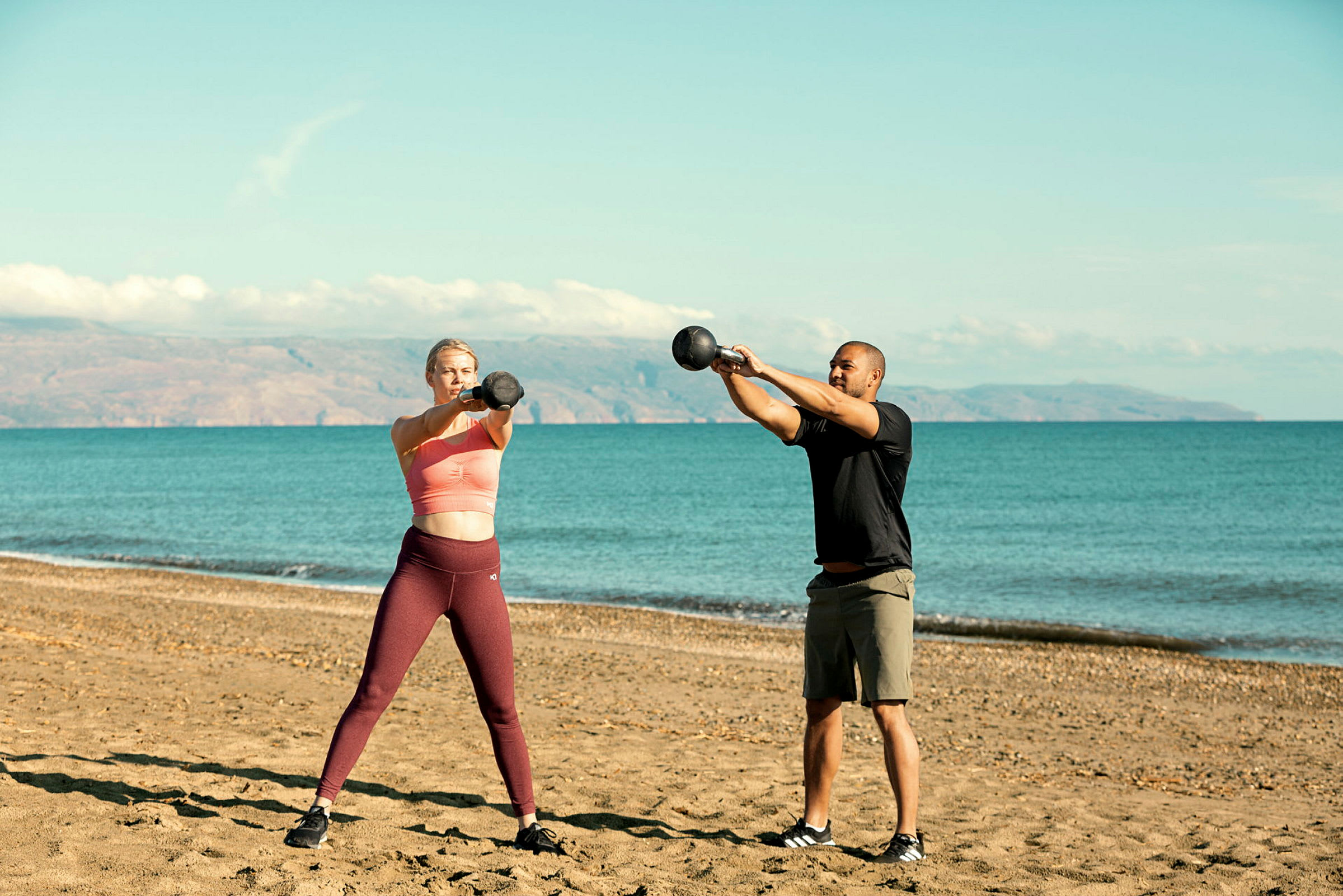To personer træner med kettlebells på en sandstrand med havet i baggrunden. (AI-genereret tekst)
