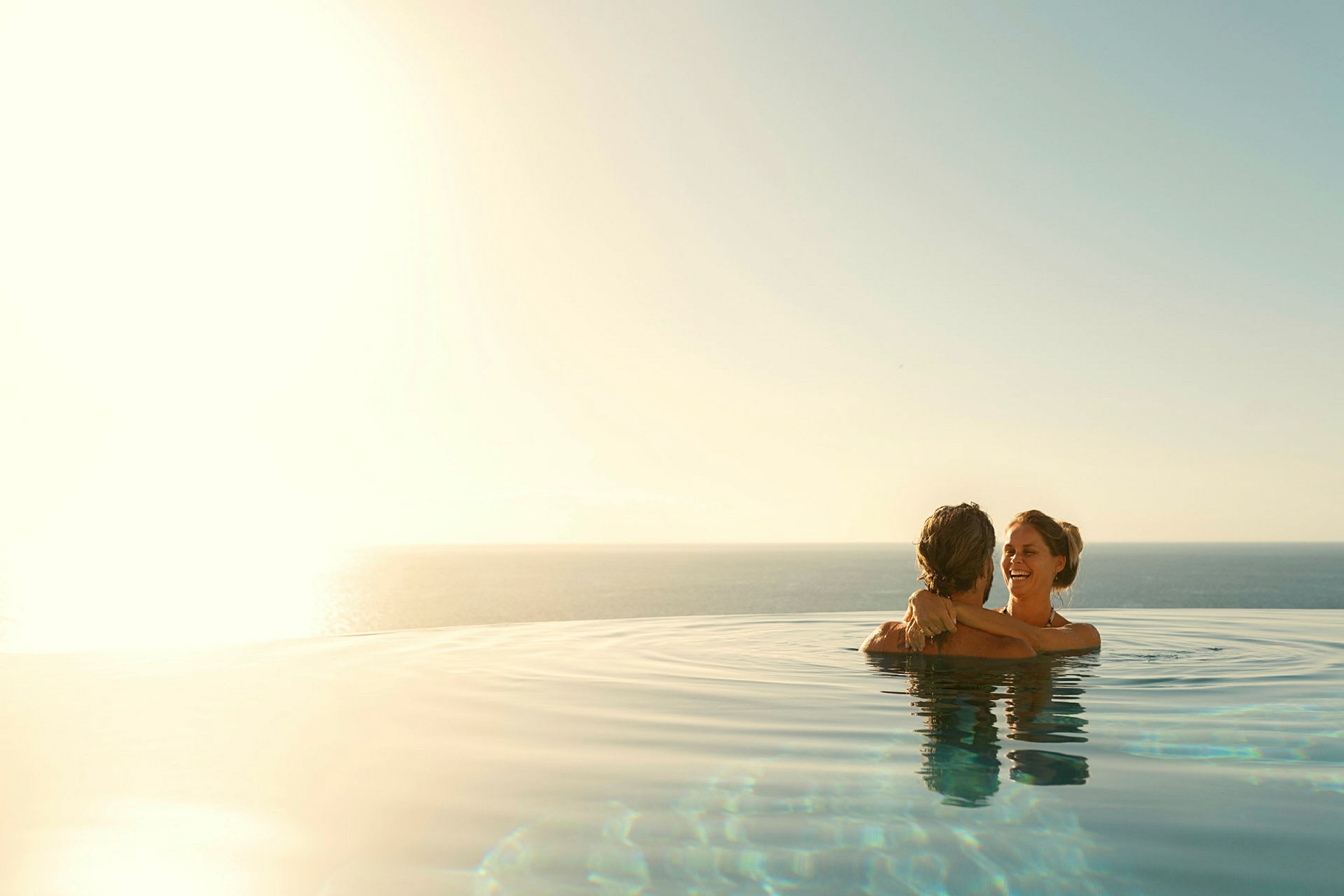 To personer holder om hinanden i en infinitypool med havudsigt under en klar himmel. (AI-genereret tekst)