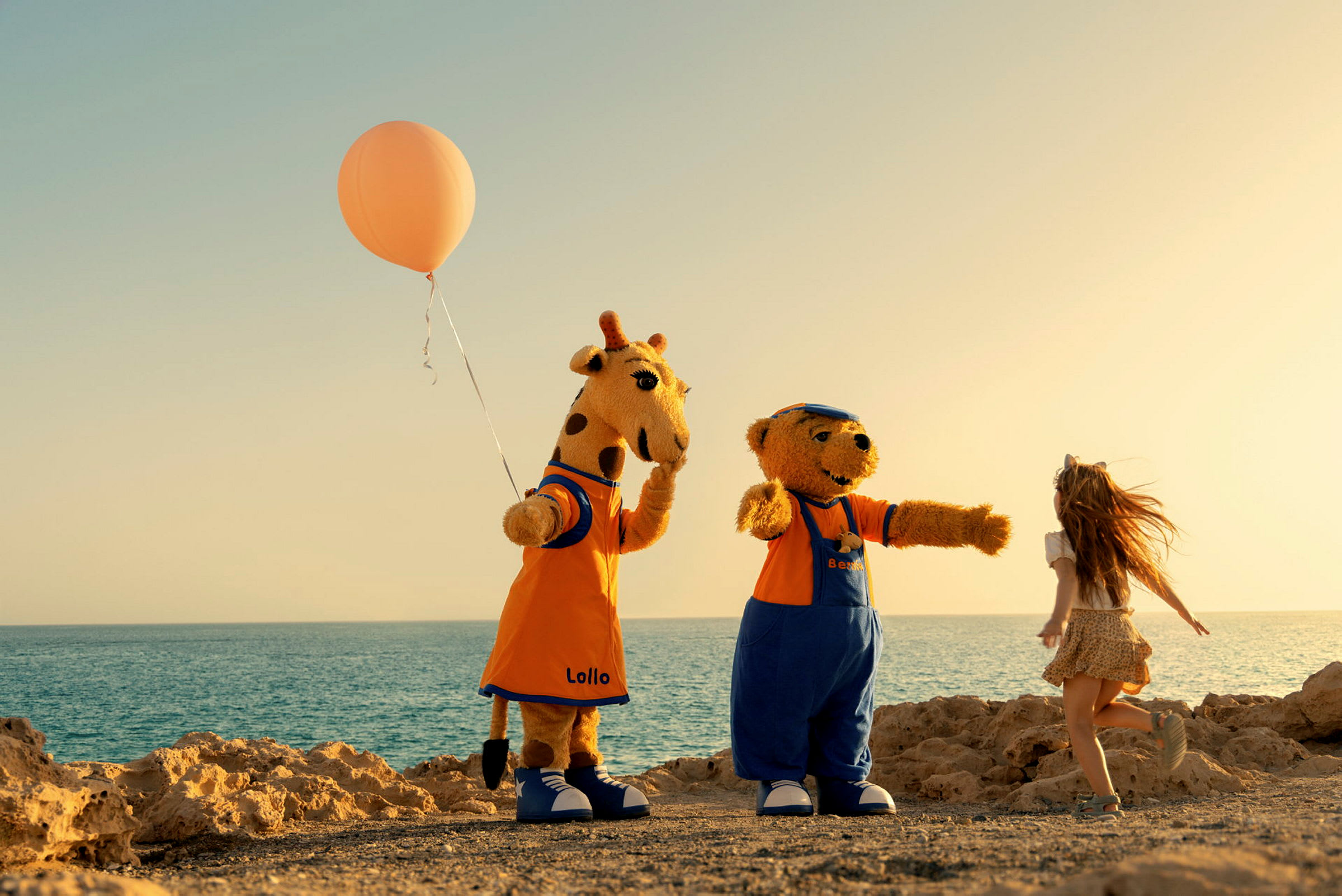To figurer klædt som en giraf og en bjørn står ved siden af et barn på en strand med havet i baggrunden. Giraffen holder en orange ballon. (AI-genereret tekst)