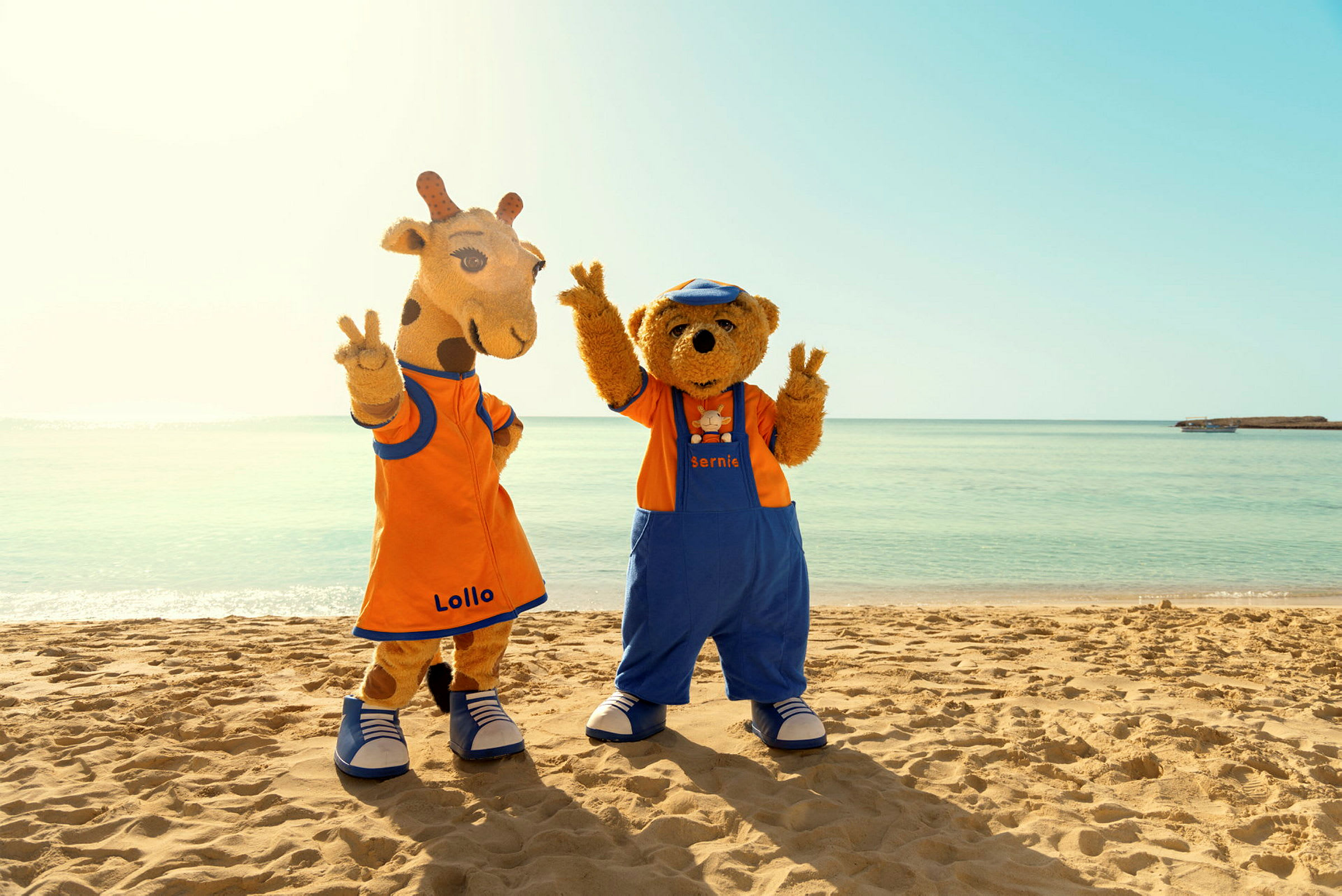 To maskotfigurer med navnene Lollo og Bernie står på en strand foran det klare hav. (AI-genereret tekst)
