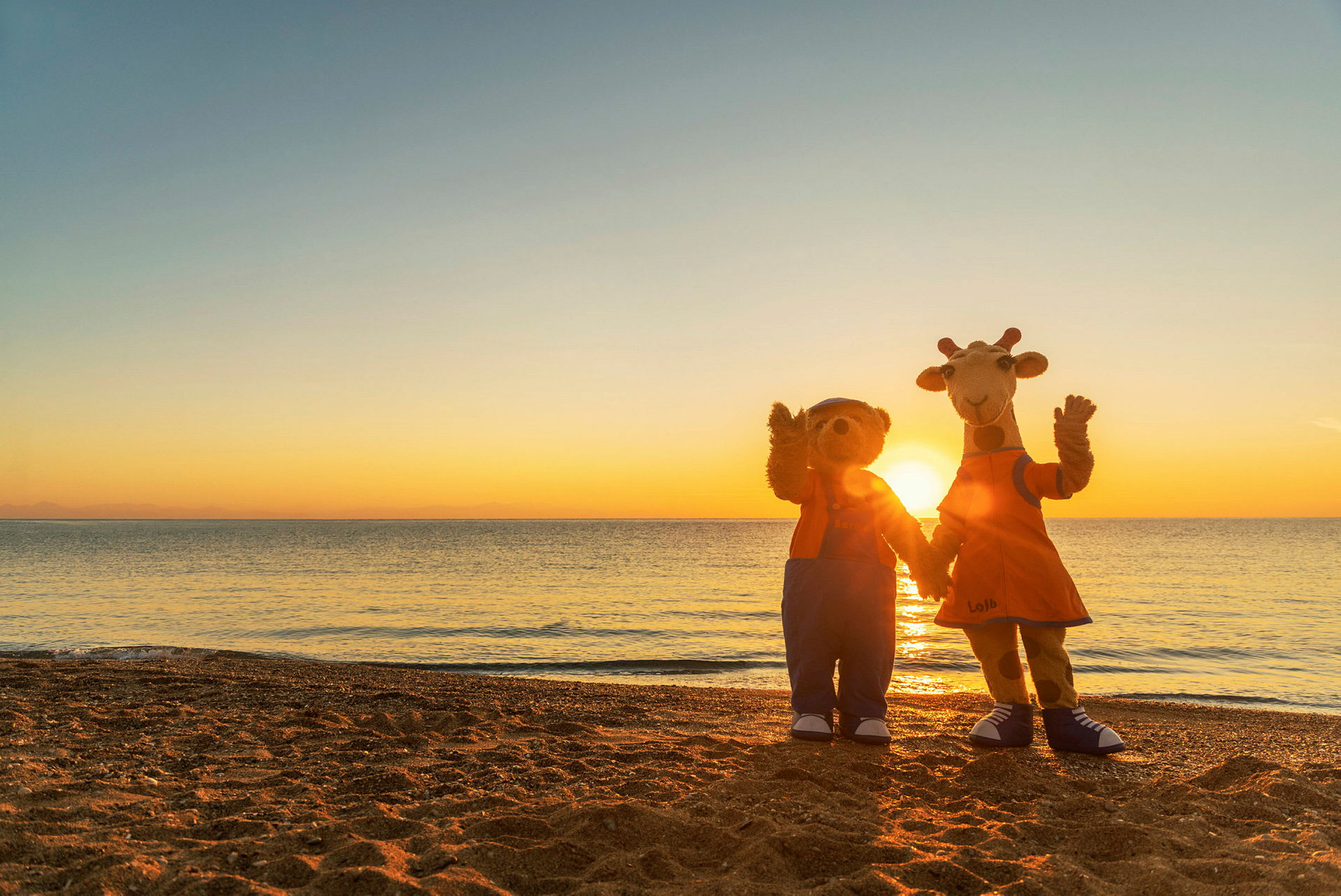 To maskotter står på en strand foran en solnedgang, vendt mod kameraet. (AI-genereret tekst)