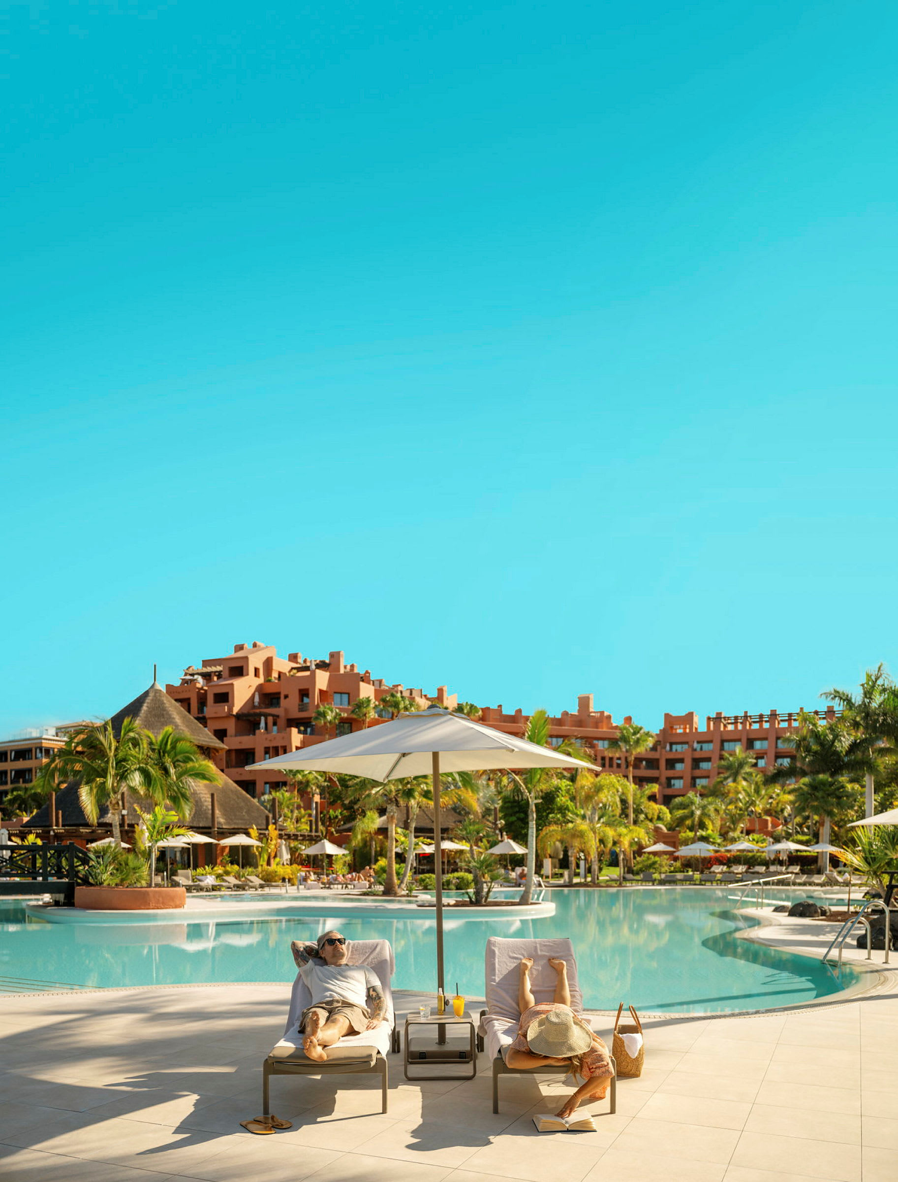 To liggestole ved kanten af en udendørs pool med en parasol, omgivet af tropiske planter og hotelbygninger i baggrunden. (AI-genereret tekst)